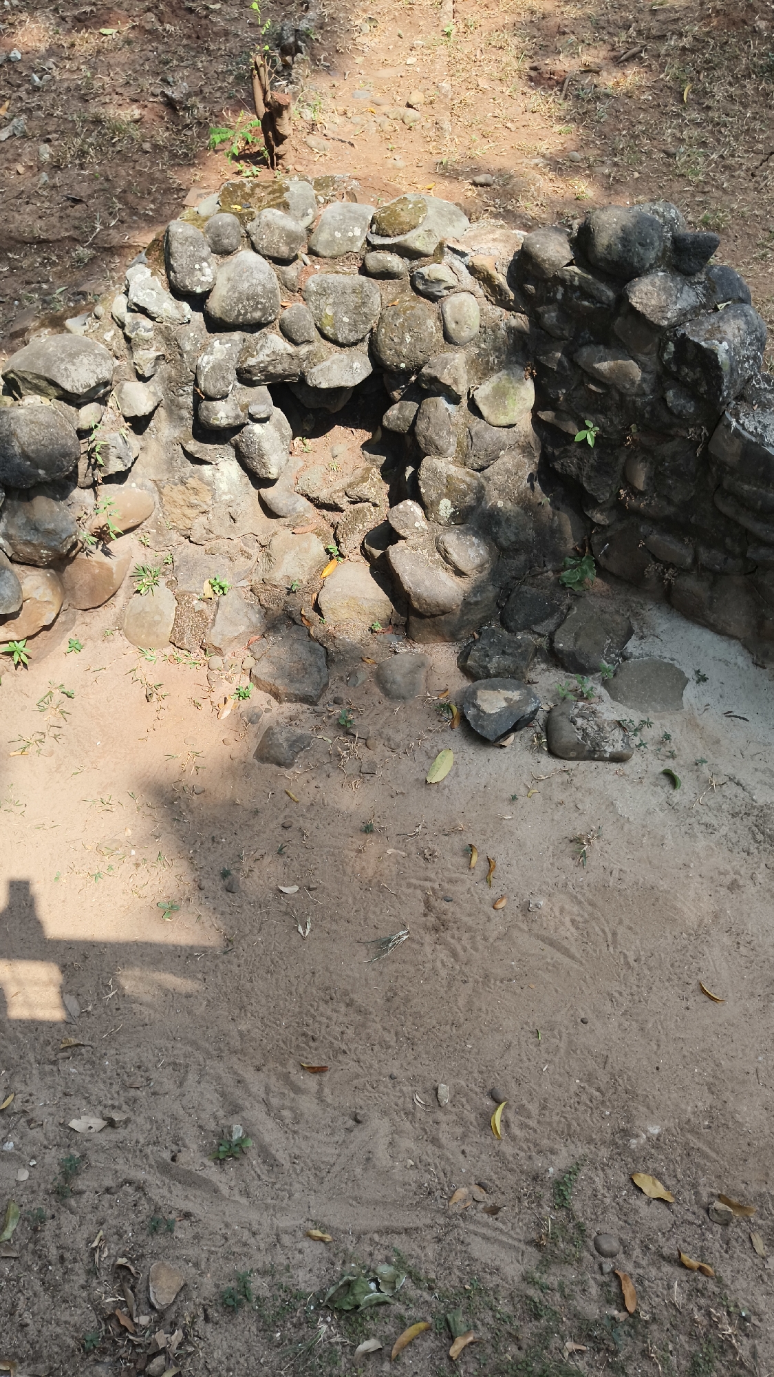 The image shows a small, partially excavated stone structure, possibly an archaeological ruin, set in a sandy area with some scattered leaves and vegetation. The structure is made of irregularly shaped, grey stones, some of which appear weathered and covered in moss. It forms a curved wall with an opening or hollow in the center. The ground surrounding the structure is sandy and uneven, with a few small green plants growing. In the upper background, dry leaves and dirt are visible. A shadow, possibly from a person or object, falls across the sandy ground in the lower left portion of the image. The overall scene suggests an outdoor location, possibly a park or a historical site. The image does not provide any specific details about the city of South Jakarta, Indonesia, or any people, actions, or interactions.