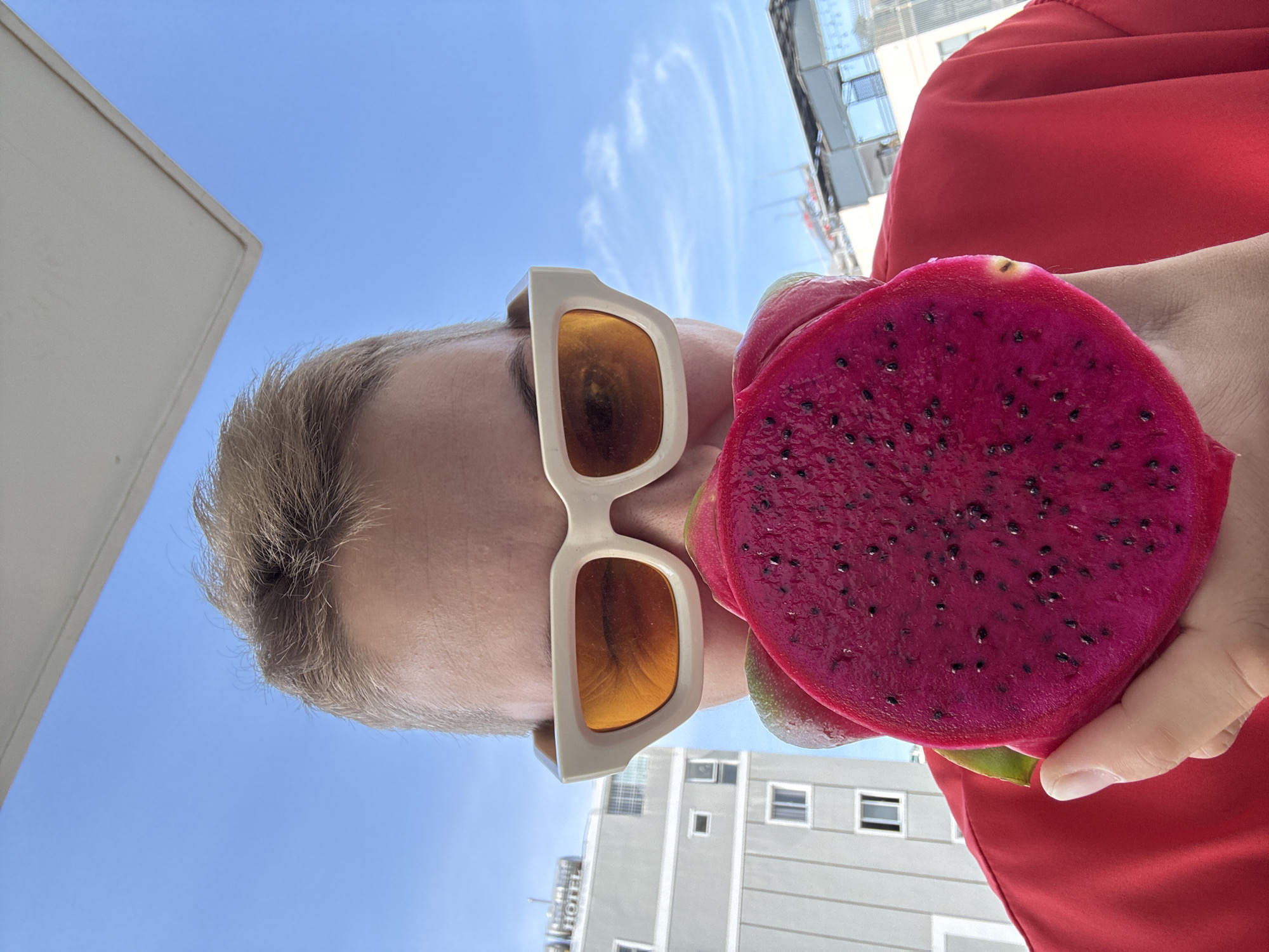 A person with short, blonde hair and stylish white-framed sunglasses with orange lenses holds a halved bright pink dragon fruit up to their face, obscuring their eyes and nose. They are wearing a vibrant red top. In the background, a grey, multi-story building with visible windows stands under a bright blue sky with scattered clouds. A partial view of a white structure, possibly a roof or awning, is visible at the top of the frame. The scene appears to be outdoors in a city environment, with elements suggestive of Nha Trang, Vietnam, such as the architectural style of the building and the clear, sunny sky typical of the region.