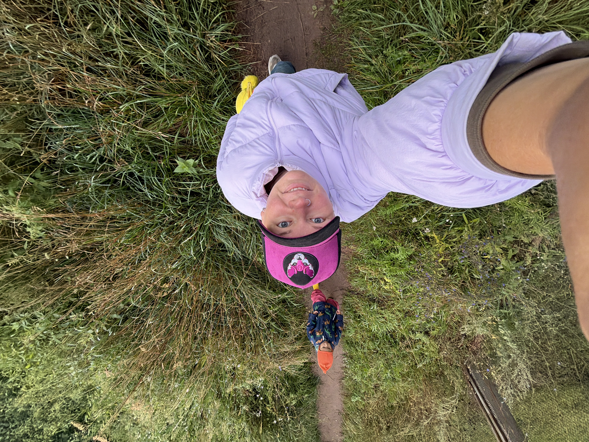 A high-angle shot shows a person in a light purple puffer jacket and a pink baseball cap, looking up at the camera with a smile. They are standing on a dirt path surrounded by tall, dry grass and green foliage. In the background, a child in a dark blue jacket with a pattern of animals and an orange hat is walking away from the camera on the same path. The child is wearing red pants. A bright yellow object, possibly a toy or backpack, is visible on the path behind the person in the puffer jacket. The environment appears to be a natural outdoor setting, possibly a park or trail within the city of Chaikovsky, Russia.