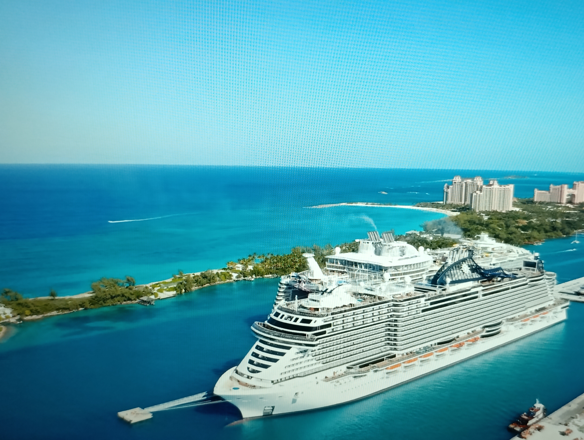 An aerial, high-angle, long shot captures a large, white cruise ship docked in a bright blue harbor. The ship, filling the lower right of the frame, is multifaceted with multiple decks, white railings, and dark-tinted windows. Several lifeboats are visible along its side. In the background, a narrow, tree-lined island separates the harbor from a wider body of turquoise water. Further in the distance, on the horizon, a cluster of tall, light-colored buildings resembling resorts or hotels are visible on land. The sky is a clear, light blue. The scene is devoid of people and any discernible action. The location is not Dubăsari town council, Moldova, but appears to be a tropical or sub-tropical coastal area.