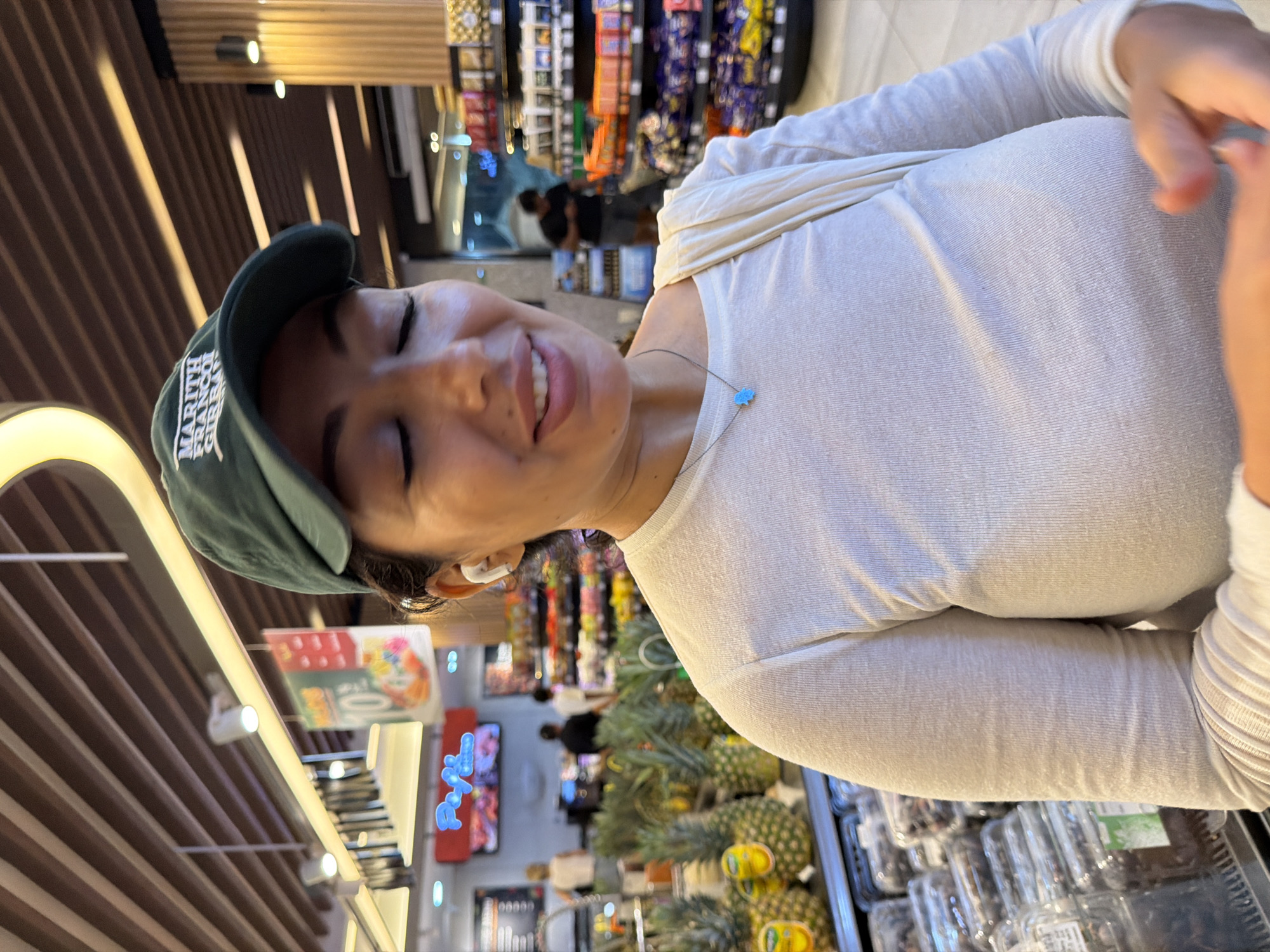 A medium shot captures a smiling woman inside a well-lit grocery store, appearing to be in North Kuta, Indonesia. She is the main subject, looking towards the camera with her eyes closed in a happy expression. She wears a dark green baseball cap, on which "MARITH FRANCOIS GIRBAUD" is partially visible. Her long-sleeved top is a light, possibly white or cream, ribbed material. A small, light blue cloud-shaped pendant hangs from a necklace around her neck, and a white earbud is visible in her right ear.
The background shows a brightly illuminated supermarket aisle with various products on shelves. To her right, a display of fresh pineapples is prominent. Further back, packaged food items in clear containers are visible. The store features a modern design with a wooden slatted ceiling and track lighting fixtures, casting distinct shadow lines. Another person is visible in the distant background, out of focus. A sign with the number "10" can be seen in the background, likely indicating a price or promotion, along with a blurry neon sign. The overall ambiance suggests a pleasant shopping experience.