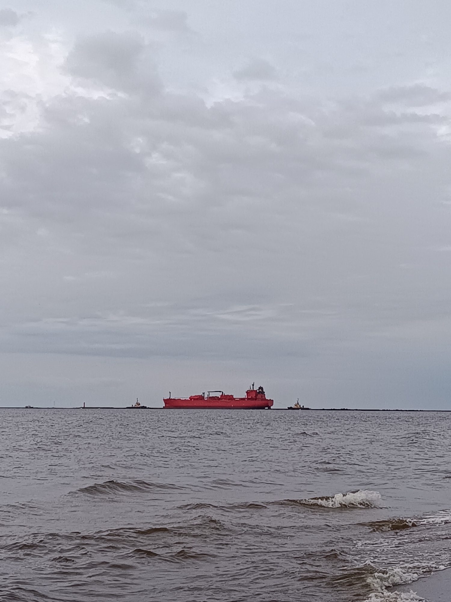 A large red oil tanker is sailing on a grey, choppy sea under a heavily overcast sky. Two tugboats are positioned alongside the tanker. In the distance, a breakwater or pier extends horizontally across the frame. The scene is likely the Gulf of Riga, near the entrance to the Port of Riga, Latvia. The water is dark and agitated, with visible waves and foam. The sky is uniformly grey, suggesting overcast or stormy weather. The overall impression is one of industrial maritime activity in a somewhat somber atmosphere.