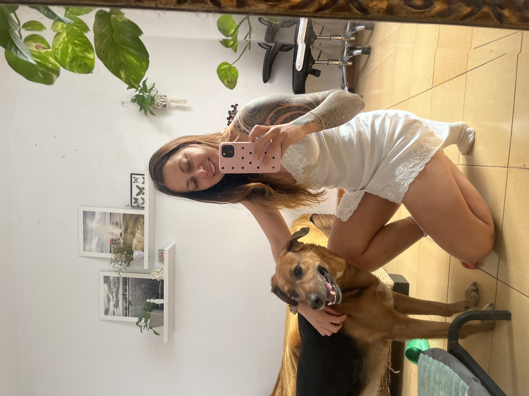 The image is a mirror selfie capturing a heartwarming interaction between a young woman and her dog in an indoor setting. The woman, with long brown hair and both arms extensively covered in intricate dark tattoos, is smiling broadly at the camera. She is dressed in a white, short slip dress or nightgown with delicate lace trim along the hem and is wearing white socks. She holds a distinctive pink smartphone, adorned with black polka dots, in her right hand to take the photo.
She is positioned kneeling or squatting beside a medium to large-sized dog, which appears to be a German Shepherd mix with brown and black fur. The woman's left arm is around the dog in an embrace. The dog, with its mouth slightly open revealing teeth and tongue, looks towards the camera with an expression of excitement or happiness.
The background reflects a brightly lit room with white walls and light beige or tan tiled flooring. Decorative elements include several green plants: a large, leafy pothos plant hangs from above the mirror, and a smaller plant is in a wall-mounted holder. Two white wall shelves display framed pictures, one a color landscape and the other a black and white landscape, along with small plant decorations. Parts of an office chair with chrome legs and a dog bed are also visible in the reflection. The scene conveys a joyful and affectionate moment, suggesting a comfortable home environment in Barcelona, Spain, during daylight hours. No discernible text is visible.