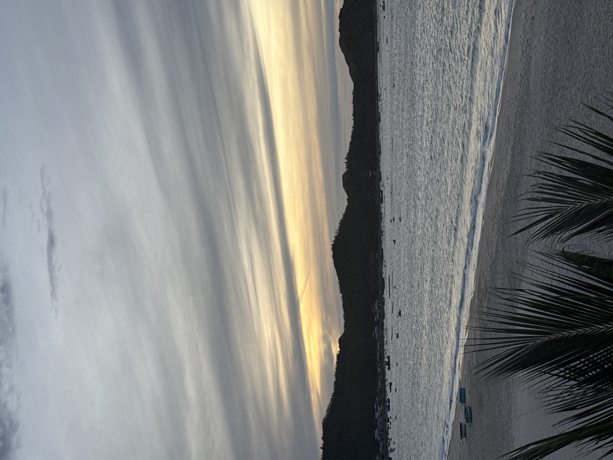 The image depicts a serene beach scene at dusk in an unknown location. The sky is filled with dramatic, horizontally striated clouds, transitioning from a pale gray at the top to a soft yellow and orange glow near the horizon, indicating the setting sun. Below the sky, a silhouetted mountain range is visible, densely covered in trees, with a small settlement of houses nestled at its base. The sea is calm, with gentle waves lapping against the sandy shore. Scattered across the water are numerous small boats, some appearing to be fishing vessels, and what looks like a raft or floating structure. In the foreground, the sandy beach is sparsely populated with three small boats pulled ashore. Palm fronds are visible at the very bottom of the frame, adding a tropical touch to the scene. The overall atmosphere is peaceful and tranquil.