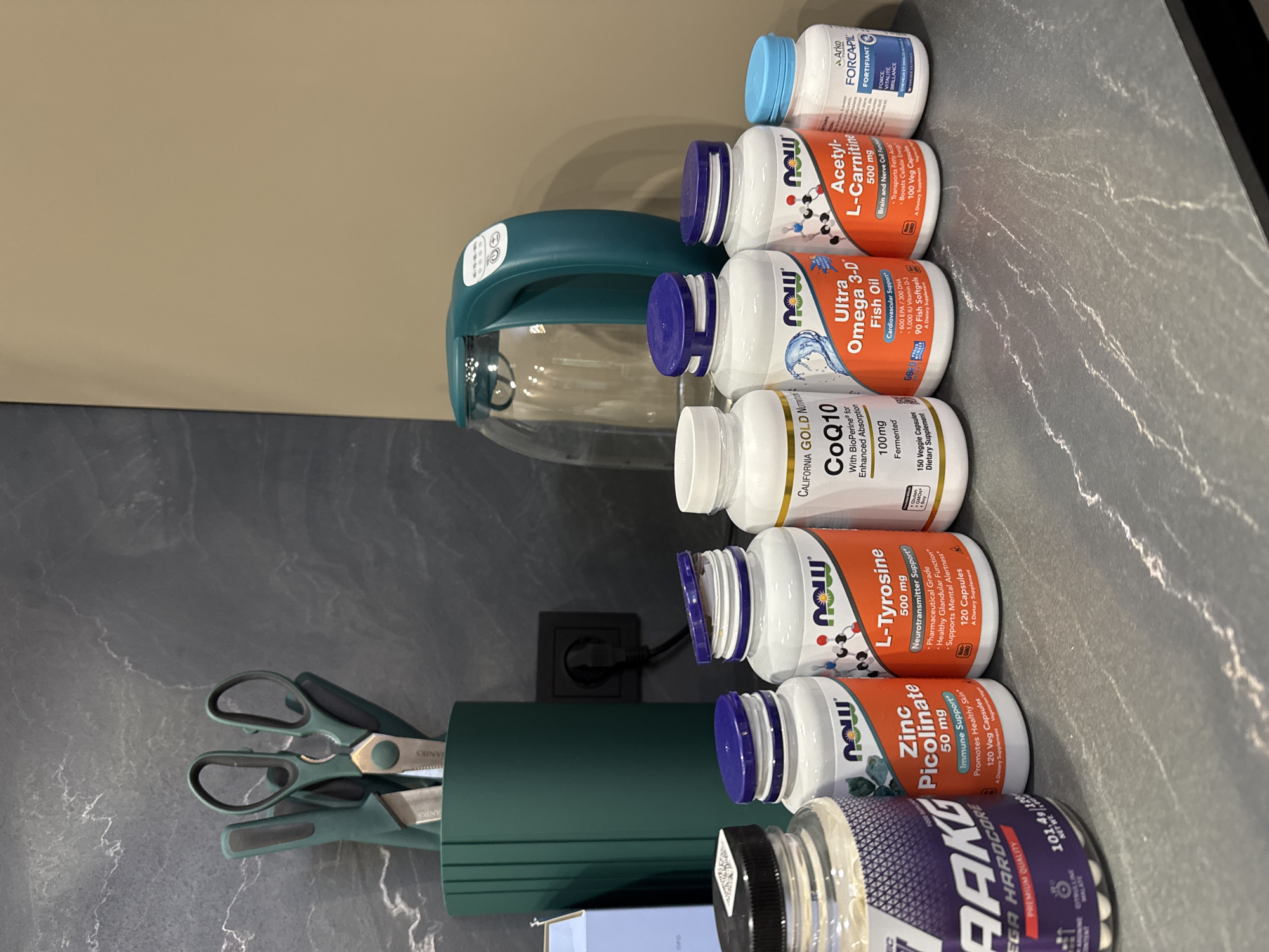 The image shows a countertop with several bottles of supplements lined up. From right to left, the supplements are "Arko FORCAPIL FORTIFIANT," "NOW Acetyl-L-Carnitine 500 mg," "NOW Ultra Omega 3-D Fish Oil," "CALIFORNIA GOLD NUTRITION CoQ10 100mg," "NOW L-Tyrosine 500 mg," "NOW Zinc Picolinate 50 mg," and an unlabeled bottle of "EGA HARDCORE" supplement.
A green water kettle and a green utensil holder containing scissors are also on the countertop. There is an electrical outlet on the wall behind the items.
The countertop is a gray, marble-like surface. The wall behind the countertop is a light brown color.