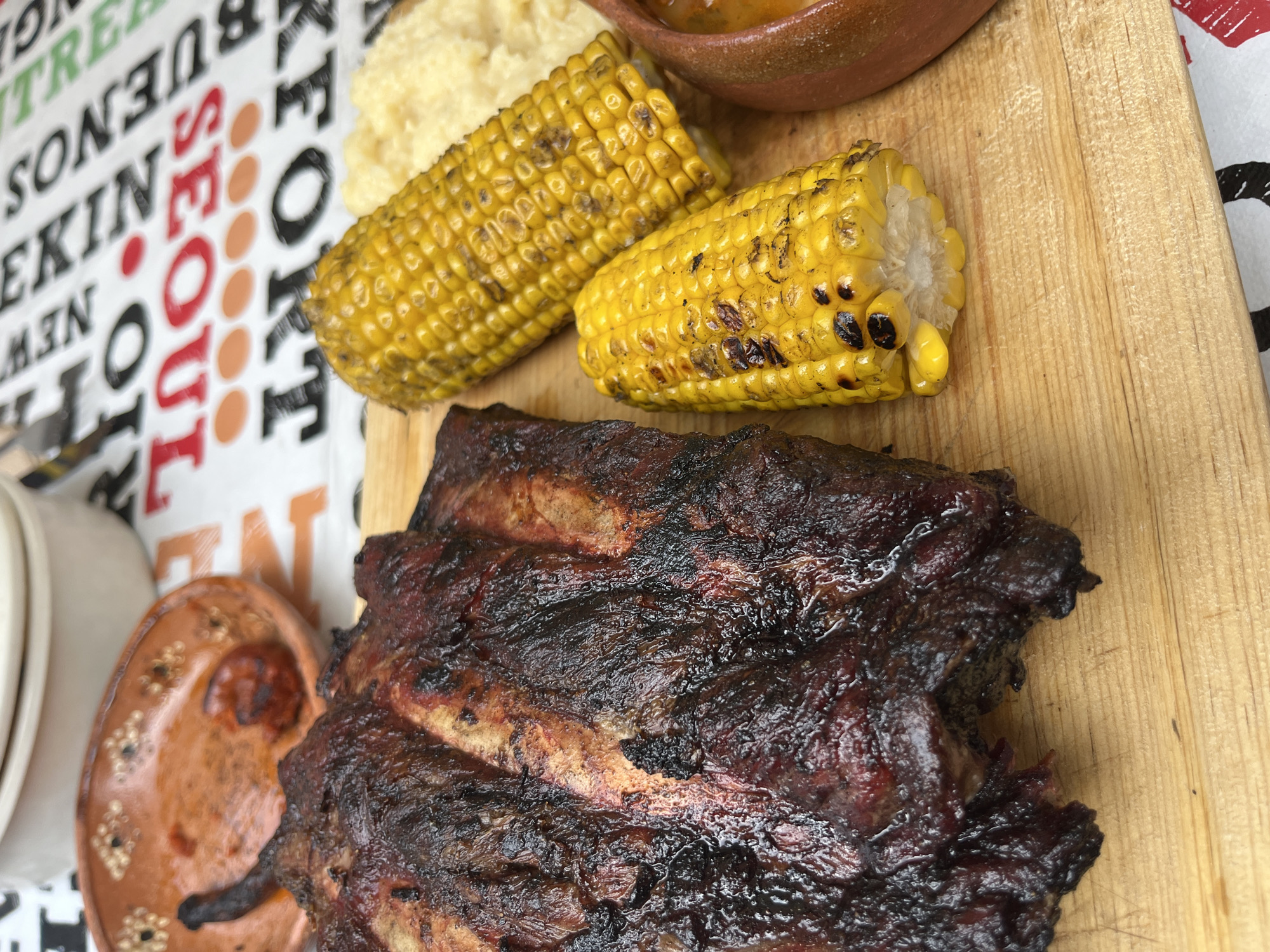 A close-up shot of a wooden board featuring food. The primary subject is a large portion of barbecued ribs, with a dark, crusty exterior. Beside the ribs are two pieces of grilled corn on the cob, slightly charred in places. A portion of what appears to be mashed potatoes or a similar side dish is also visible. Additionally, there are small ceramic bowls containing sauce. The background consists of a table covering with various city names, including "SEOUL" in red letters and "NEW YORK" and "PEKIN" in black. The location, San Francisco Totimehuacán, Mexico, cannot be determined from the image content.