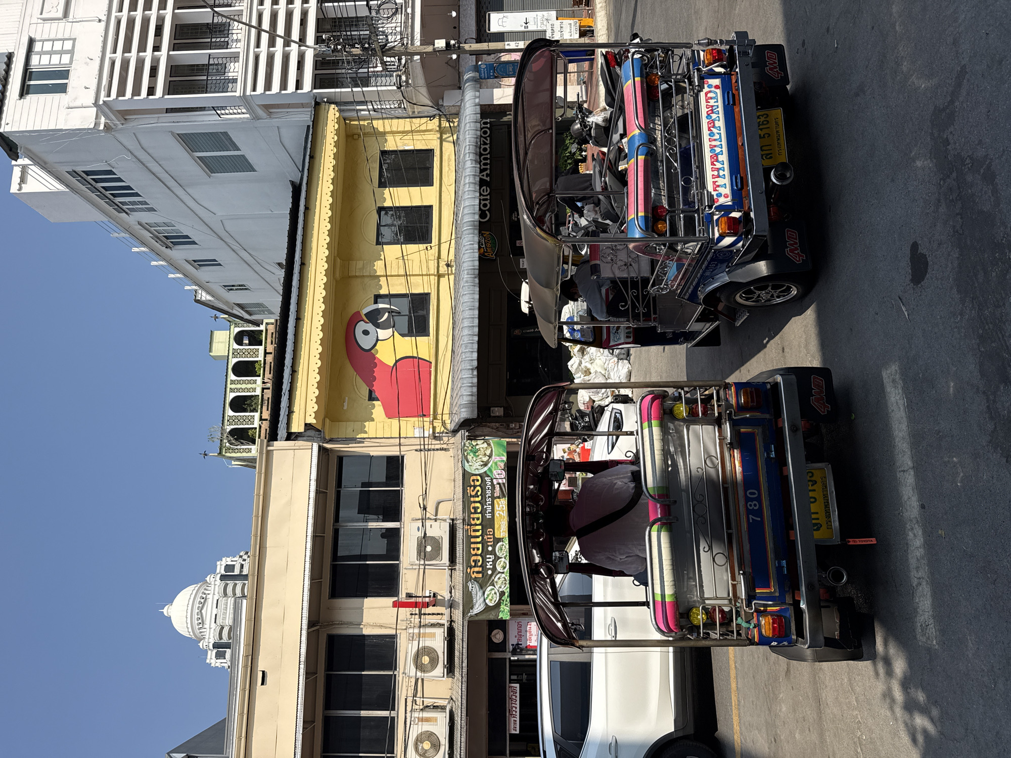 The image captures an urban street scene in Bangkok, Thailand, under a clear blue sky. Two multi-colored Tuk-Tuks are positioned in the foreground, viewed from their rear. The left Tuk-Tuk is predominantly blue with pink and green accents, displaying "780" and a Thai license plate "ฒค 0153 Bangkok". Its driver is visible from the back, wearing a light-colored shirt. A white SUV is partially visible behind and to the left of this vehicle. The right Tuk-Tuk, similarly blue with pink and red accents, features "THAILAND" on its rear and a Thai license plate "สค 5163 Bangkok". Two motorcycles are stored in the rear compartment of this Tuk-Tuk. Both Tuk-Tuks have "4WD" mudflaps.
In the midground, a yellow building prominently displays a large red and yellow parrot head mural on its upper facade. Below, a grey awning extends over the entrance to "Cafe Amazon". To the immediate left, a beige two-story building features three external air conditioning units and a banner advertising "ก๋วยเตี๋ยวเรือ" (Boat Noodles) for "25 ฿". Further right, a series of multi-story white buildings with varied window designs and balconies rise, intersected by utility poles dense with tangled power lines. In the distant background, a large, white, multi-tiered building crowned with a distinctive dome is visible against the horizon. The asphalt road surface exhibits faint yellow markings, and pronounced shadows indicate direct overhead sunlight.