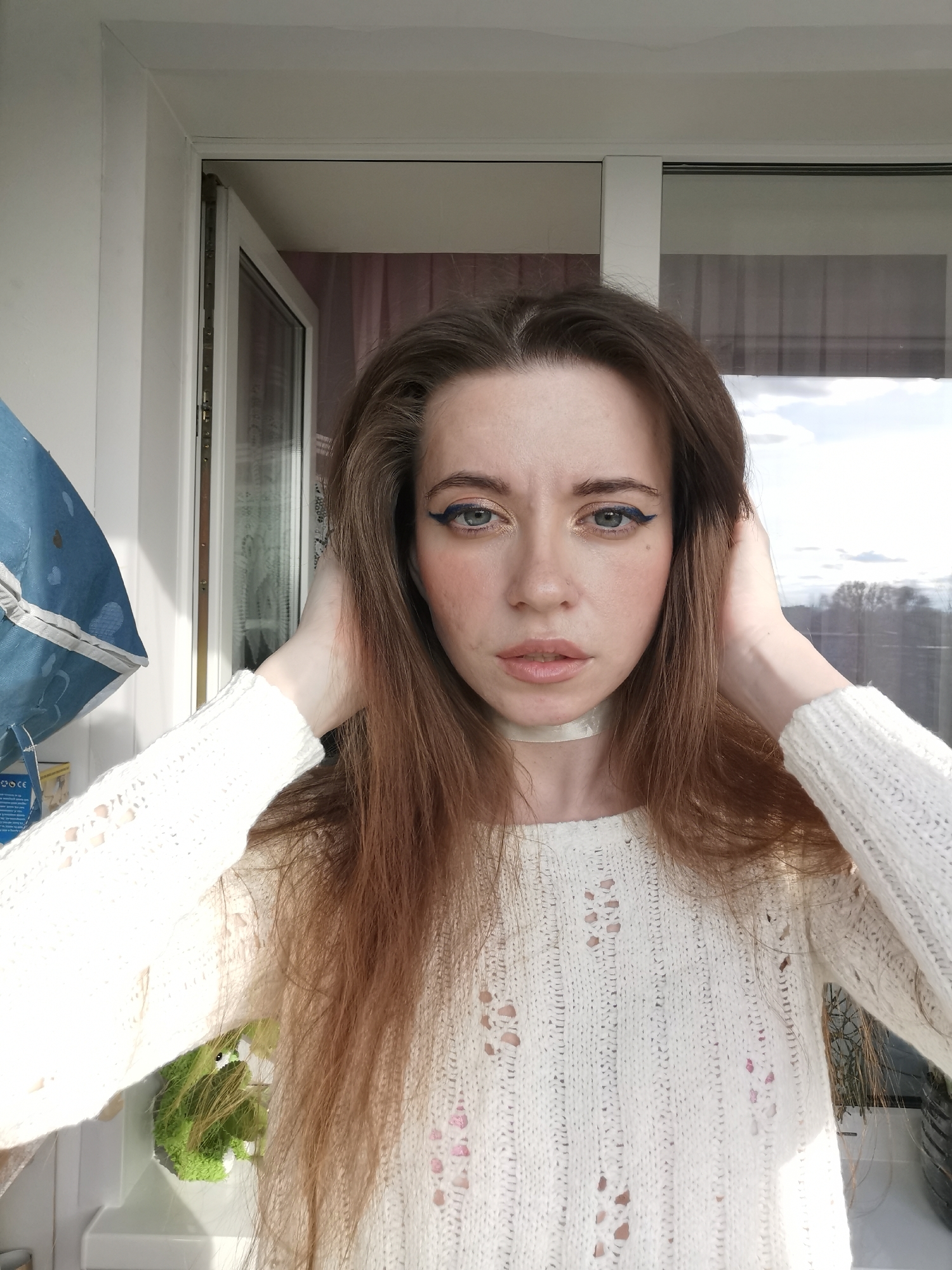 A young woman with long brown hair and light skin stands in front of an open doorway. She is wearing a white sweater and a light-colored choker. Her hands are raised to her head, fingers in her hair. She has eyeliner and blush on her face. Behind her, a door frame opens to an outdoor area with a view of trees and a cloudy sky. The location appears to be indoors.