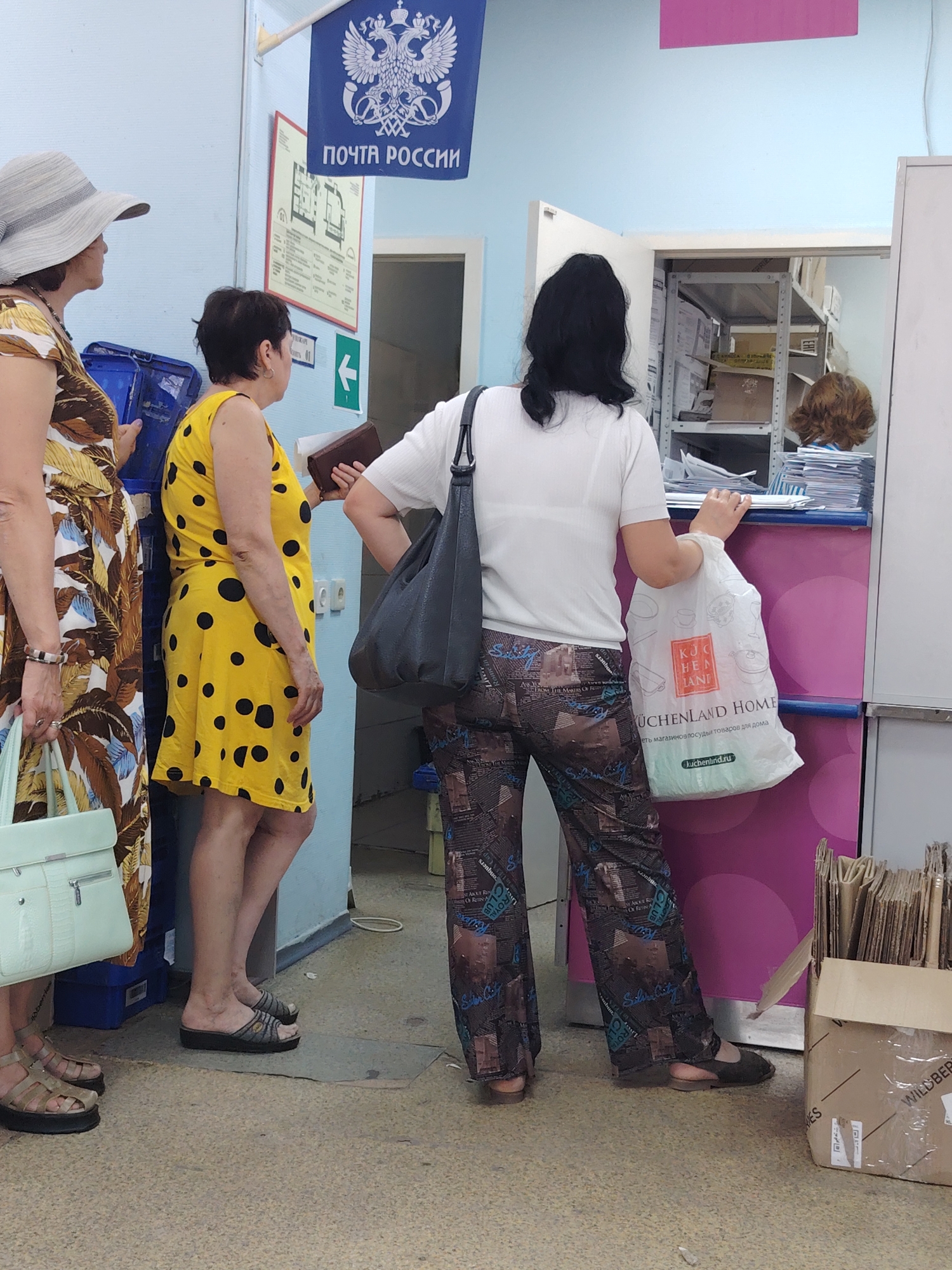 In this image, three women are standing in a post office in Rostov-on-Don, Russia. The woman on the far left wears a wide-brimmed straw hat and a floral print dress. She carries a light-colored handbag. Next to her, a woman in a yellow polka-dot dress holds a brown wallet. The woman on the right, facing away from the camera, is dressed in a white t-shirt and patterned trousers. She holds a white plastic shopping bag with the text "KÜCHENLAND HOME" visible.
The post office setting is indicated by a blue flag with the Russian double-headed eagle emblem and the text "ПОЧТА РОССИИ" (Post of Russia). A directional arrow points to the left. Shelves filled with boxes and papers are visible behind the counter, where another person, partially obscured, appears to be working. The overall atmosphere suggests a typical day at a Russian post office, with people waiting to conduct their business. The lighting indicates it is daytime.
