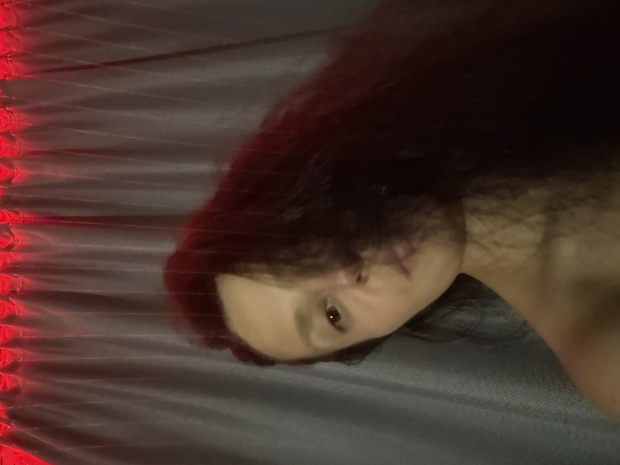 An intimately framed image, taken at an angle, depicts a person, likely female, with long, dark, wavy hair. Only a portion of her face is visible, including one eye with a direct, steady gaze, part of her nose, and her lips, suggesting a neutral or contemplative expression. Her bare shoulder or neck is also visible on the right side of the frame.
The setting appears to be indoors, characterized by dim lighting and a prominent red glow emanating from a textured, pleated, or ruffled fabric element on the left side of the image. This red light casts a warm hue across the scene. The person is resting on a grey, subtly textured fabric surface, possibly a bedsheet or upholstery, which dominates the background. The low light and artificial illumination imply the image was taken during the evening or night. There are no other identifiable objects or visible text. The overall atmosphere is quiet and personal.