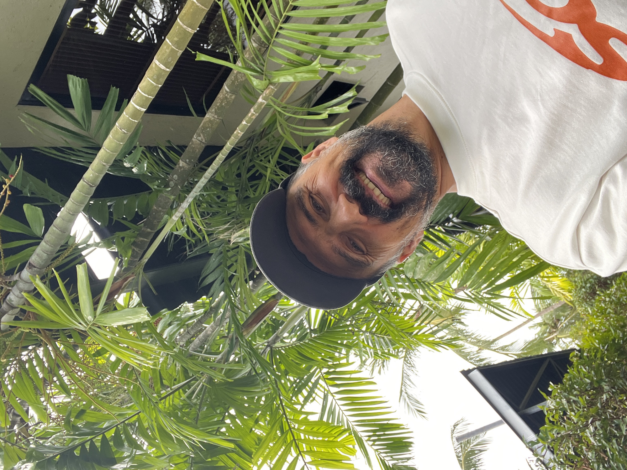 A man wearing a white T-shirt and a cap is smiling and looking at the camera. The image is taken from a low angle, with palm trees and the underside of a building visible in the background. The location appears to be an outdoor area in บ้านบางรักษ์, Thailand, based on the presence of tropical vegetation and the architecture, which are consistent with the region.
