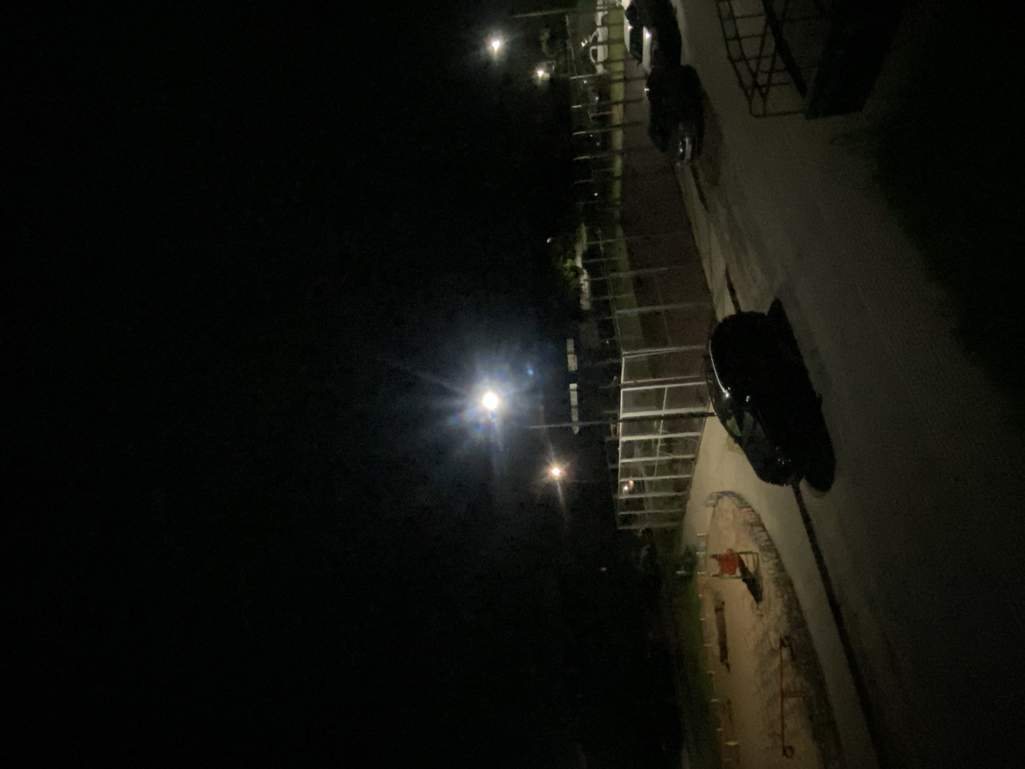 An outdoor nighttime scene shows a playground with sparse equipment under dim lighting. A black sedan is parked on a paved surface adjacent to the playground. Further down the paved surface, two more vehicles are parked, one appearing to be a light-colored sedan and the other a darker vehicle. Beyond the parked cars, a fenced sports court is visible, illuminated by overhead streetlights. The overall environment appears to be a public space in a city.