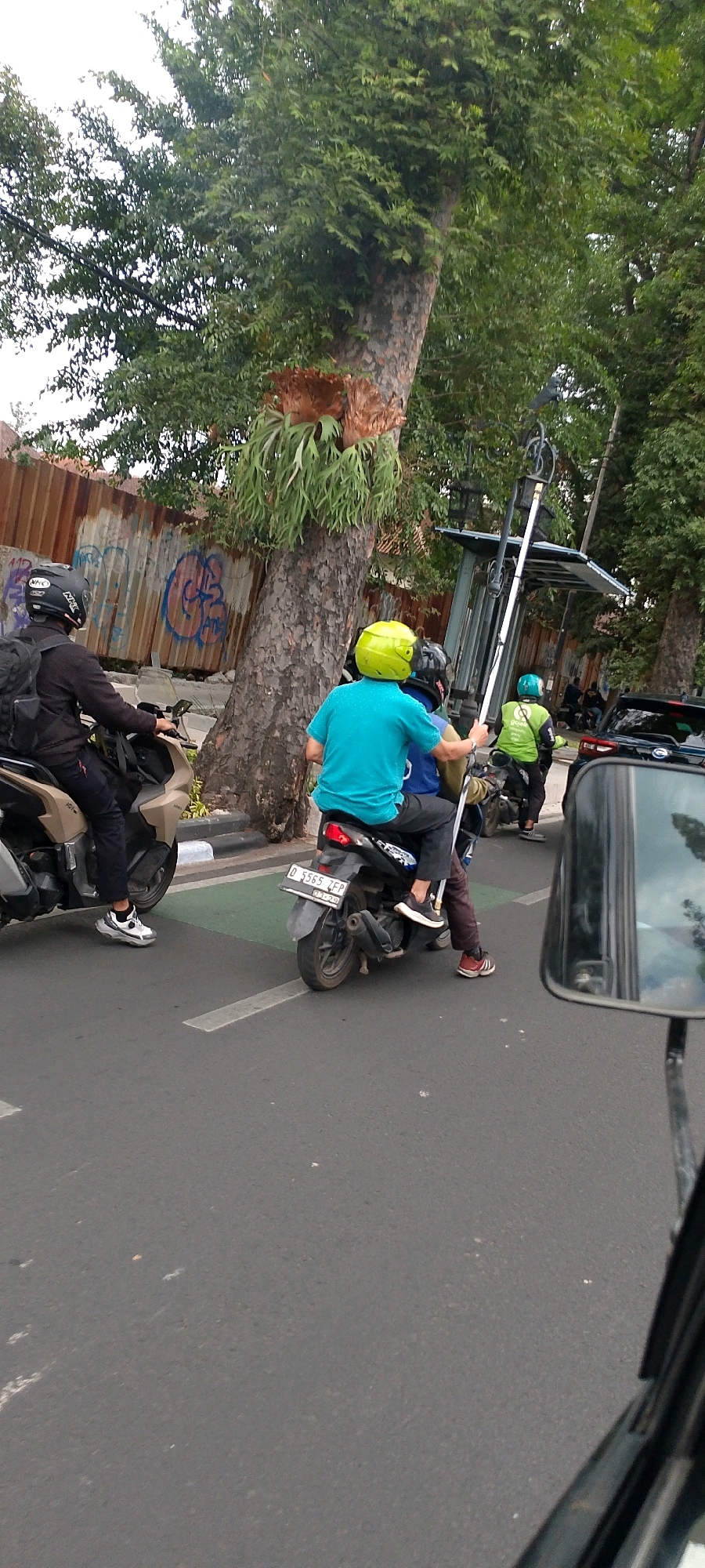 The image captures a street scene in Bandung City, Indonesia, likely during daytime due to the visible light. A person wearing a black helmet and backpack is riding a beige scooter on the left side of the road. Further ahead, two individuals are on a black scooter. The person in front wears a bright turquoise shirt and a neon yellow helmet. The person behind them is partially obscured, wearing a green jacket with "Gojek" branding and a green helmet. They appear to be holding a long pole or equipment. The scooter has a license plate that reads "D 5565 IFP." In the background, a corrugated metal fence with graffiti and a large tree with a staghorn fern growing on it are visible. A bus stop shelter can be seen to the right of the tree. Several other vehicles, including a black car, are present further down the road. The overall setting suggests an urban environment with moderate traffic. The perspective is from another vehicle, likely a motorcycle, as indicated by the reflection in the side mirror on the right side of the frame.