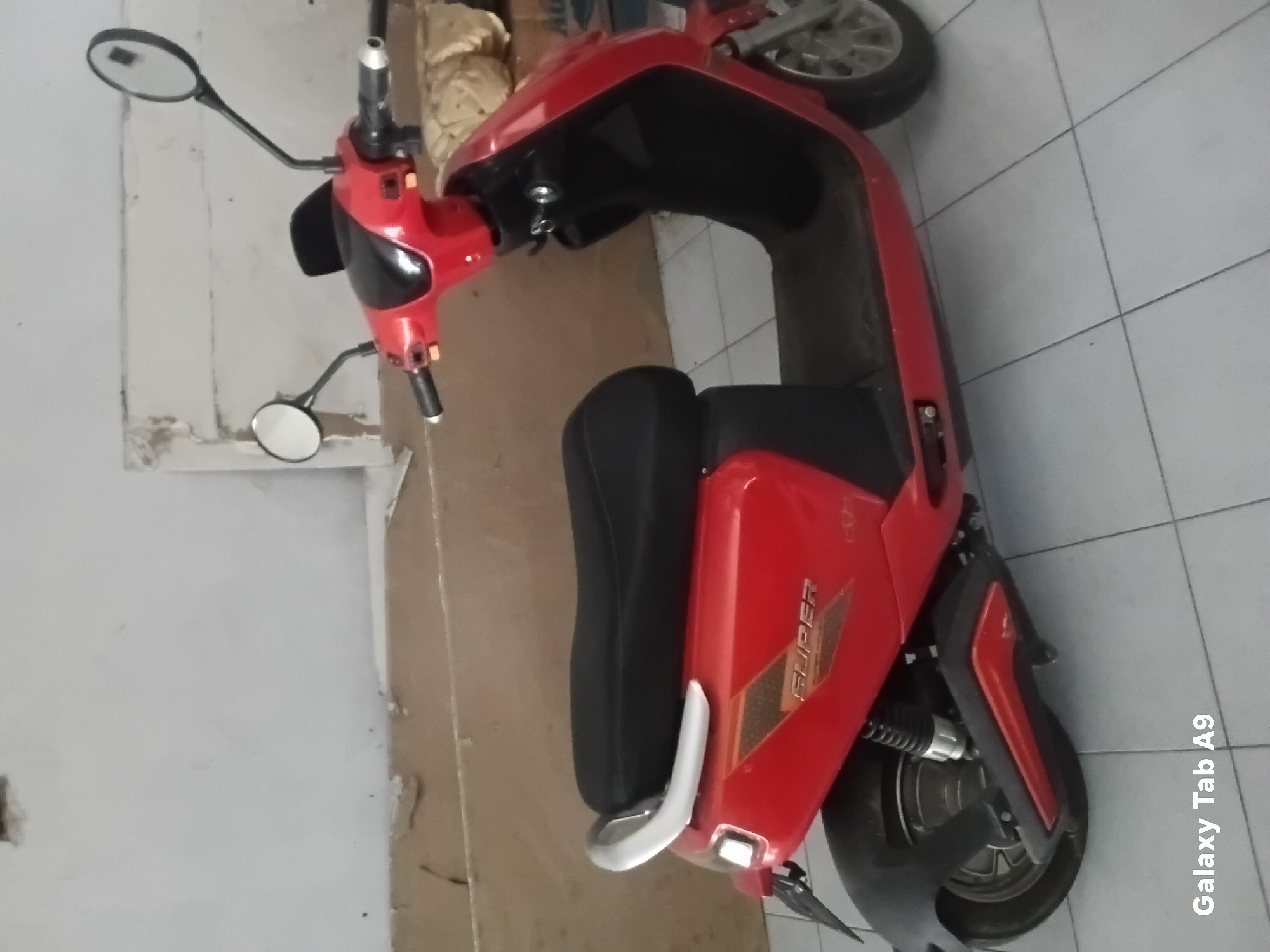 A red electric scooter, identified by the "SUPER" logo on its side, is parked indoors on a tiled floor. The scooter is positioned at an angle, facing towards the right of the frame. It features a black seat, silver handlebars with black grips, and two rearview mirrors. The background consists of a white wall with some discoloration and what appears to be a large, brown cardboard box or partition. To the bottom left of the image, the text "Galaxy Tab A9" is visible, indicating the device used to capture the photograph. The scene suggests an indoor storage or garage environment within the city of Kelutan, Indonesia.