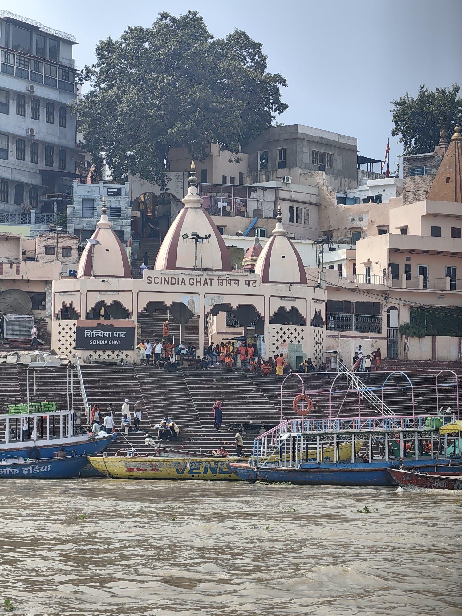 The image shows a view of Scindia Ghat in Varanasi, India, from the Ganges River. In the foreground, there are several boats moored on the water, some of which are decorated with colorful patterns. Behind the boats are the ghat steps, which are lined with people sitting and standing. At the top of the steps is a temple complex with prominent domes and intricate architecture. The temple is adorned with religious inscriptions in both Hindi and English. The surrounding buildings are multi-story structures with balconies and windows, indicating an urban environment. The sky is clear and blue, suggesting a sunny day. The overall scene depicts a bustling religious and cultural hub.