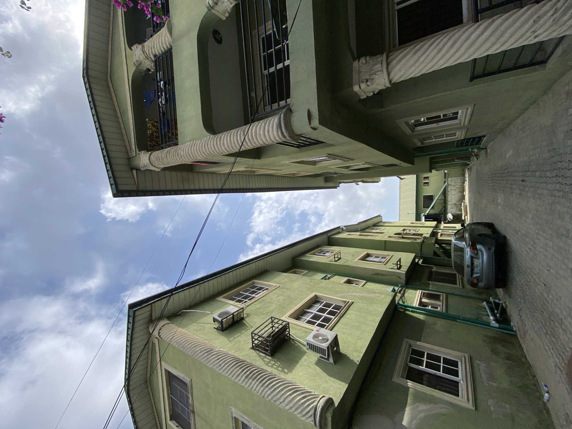 The image depicts two green buildings, likely apartments, facing each other across a narrow street in Ado, Nigeria. The buildings feature white-framed windows, air conditioning units, and decorative architectural elements. A gray car is parked on the street between the buildings. The sky above is visible with clouds. No people are visible in the image.
