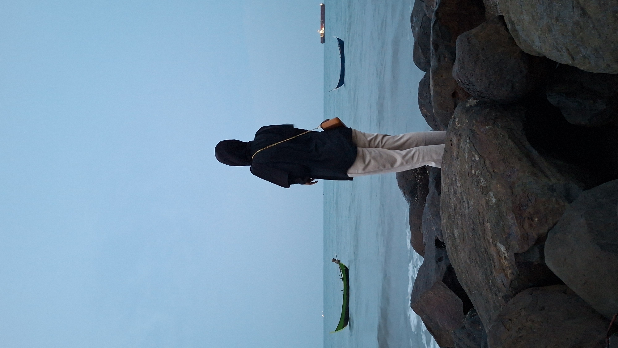 A person wearing a black hijab and light-colored pants stands on a rocky shore facing the ocean in Lhokseumawe, Indonesia. A small, green boat is anchored in the distance to the left, and a dark blue boat is closer on the right. Further out on the horizon, a large cargo ship is visible. The sky is a soft blue, and the water is calm. The rocks in the foreground are large and dark.
