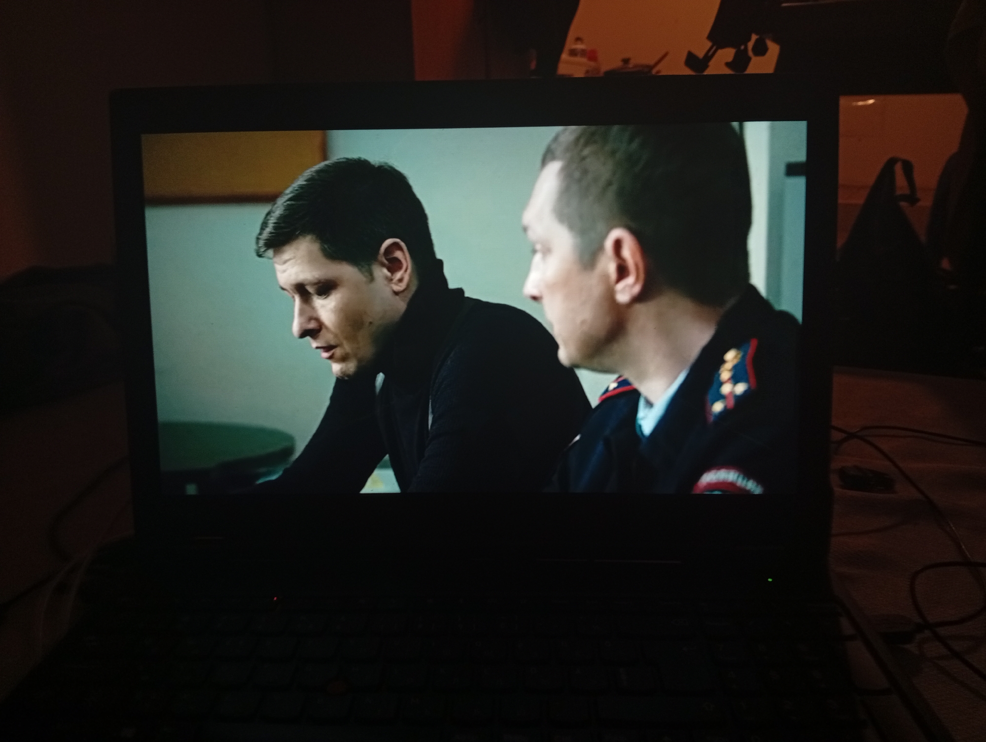 The image displays a laptop screen showing a scene from a movie or TV show. Two men are visible in the frame. The man on the left has dark hair and is wearing a black turtleneck. He is looking downwards, with his mouth slightly open as if speaking. The man on the right has lighter hair and is wearing a dark uniform with red and gold insignia on the collar and shoulder. He is looking towards the man on the left. The background on the screen is a muted teal color with a suggestion of a shelf or frame in the upper left. The laptop itself is dark and appears to be on a table or desk. Wires are visible on the right side of the laptop. The environment outside the laptop screen is dimly lit. There is no information in the image to suggest any connection to Mladá Boleslav, Czechia.