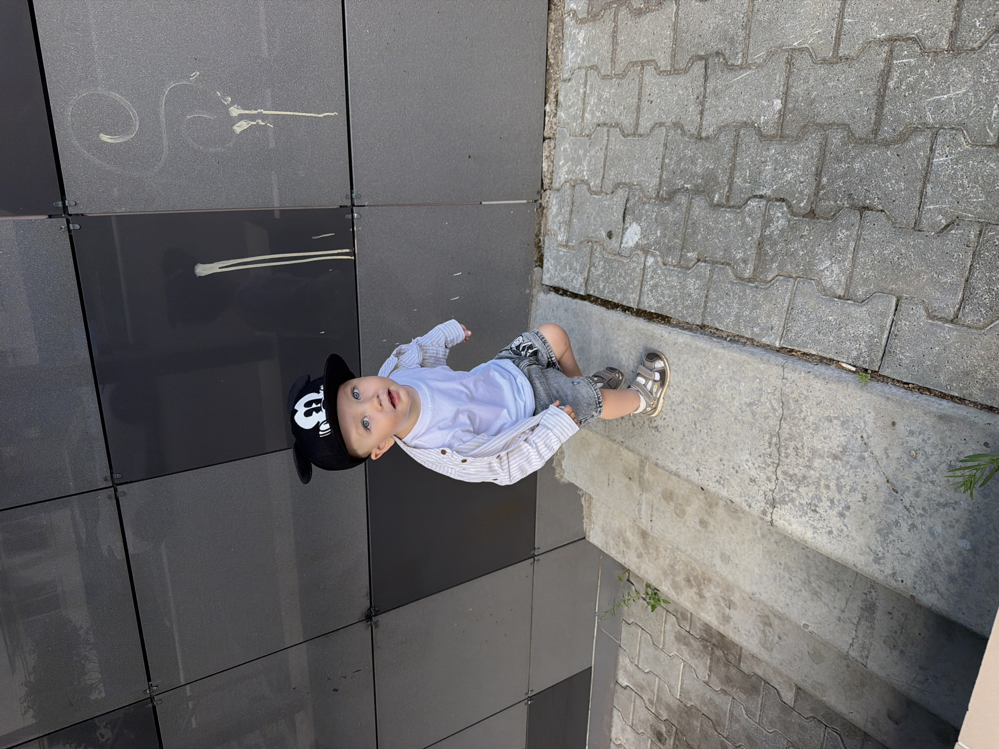 A young boy wearing a Mickey Mouse hat, a white t-shirt, a light-colored striped button-down shirt, and grey shorts is standing on a concrete stairway. He is looking up and to his right. The stairway is adjacent to a paved area with interlocked grey pavers. Behind the stairway is a modern building with a dark grey tiled facade, some of which has white markings. The scene appears to be outdoors in an urban setting, possibly a city like Chaikovsky, Russia, given the architecture and paving style.