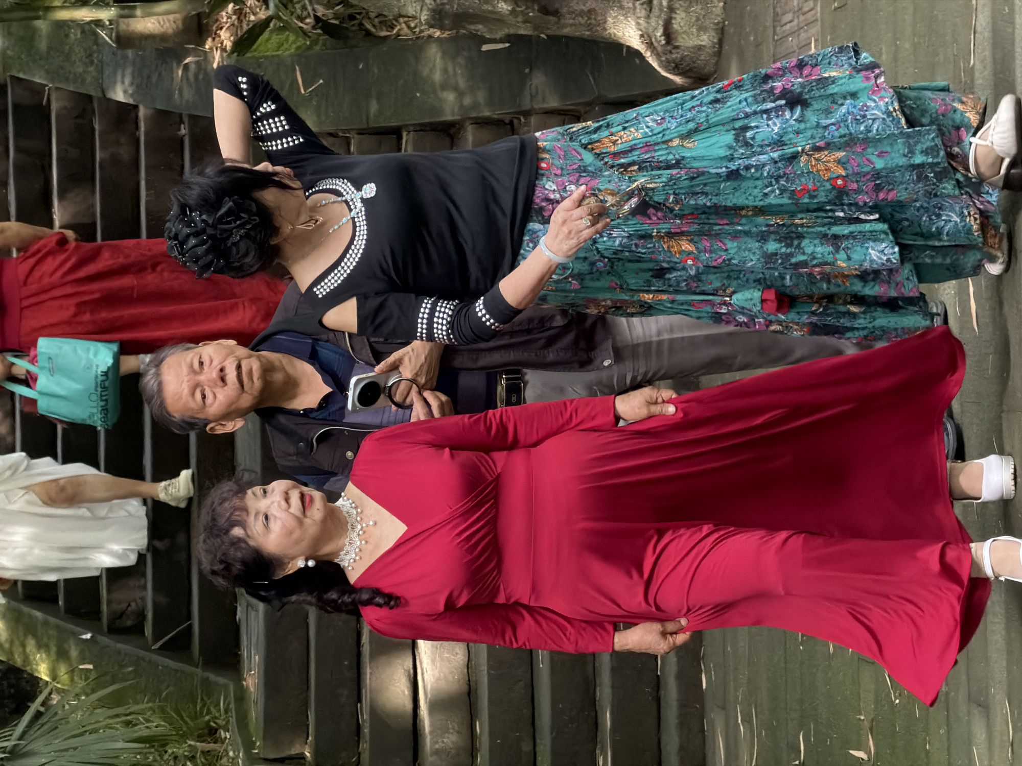 In Yuzhong District, China, three individuals are captured on stone steps amidst lush greenery. The foremost subject, a woman in a vibrant red full-length dress with a V-neck and cinched waist, stands at the bottom left of the frame. She wears a pearl and crystal necklace and white mary jane heels. To her right, a man in a dark jacket over a blue shirt holds a smartphone in his right hand. Behind him and to the right, a woman with elaborate dark hair adorned with black flowers wears a black off-the-shoulder top with crystal embellishments on the sleeves and neckline, paired with a flowing, multi-colored patterned skirt in shades of teal, pink, and orange. She is also wearing a statement necklace with a blue stone pendant and white sandals. In the background, on the steps above, another person in a flowing white garment and white sneakers is visible, as is a person in a red dress and a teal tote bag. The environment suggests an outdoor setting, possibly a park or a historical site, with stone pathways and abundant foliage.