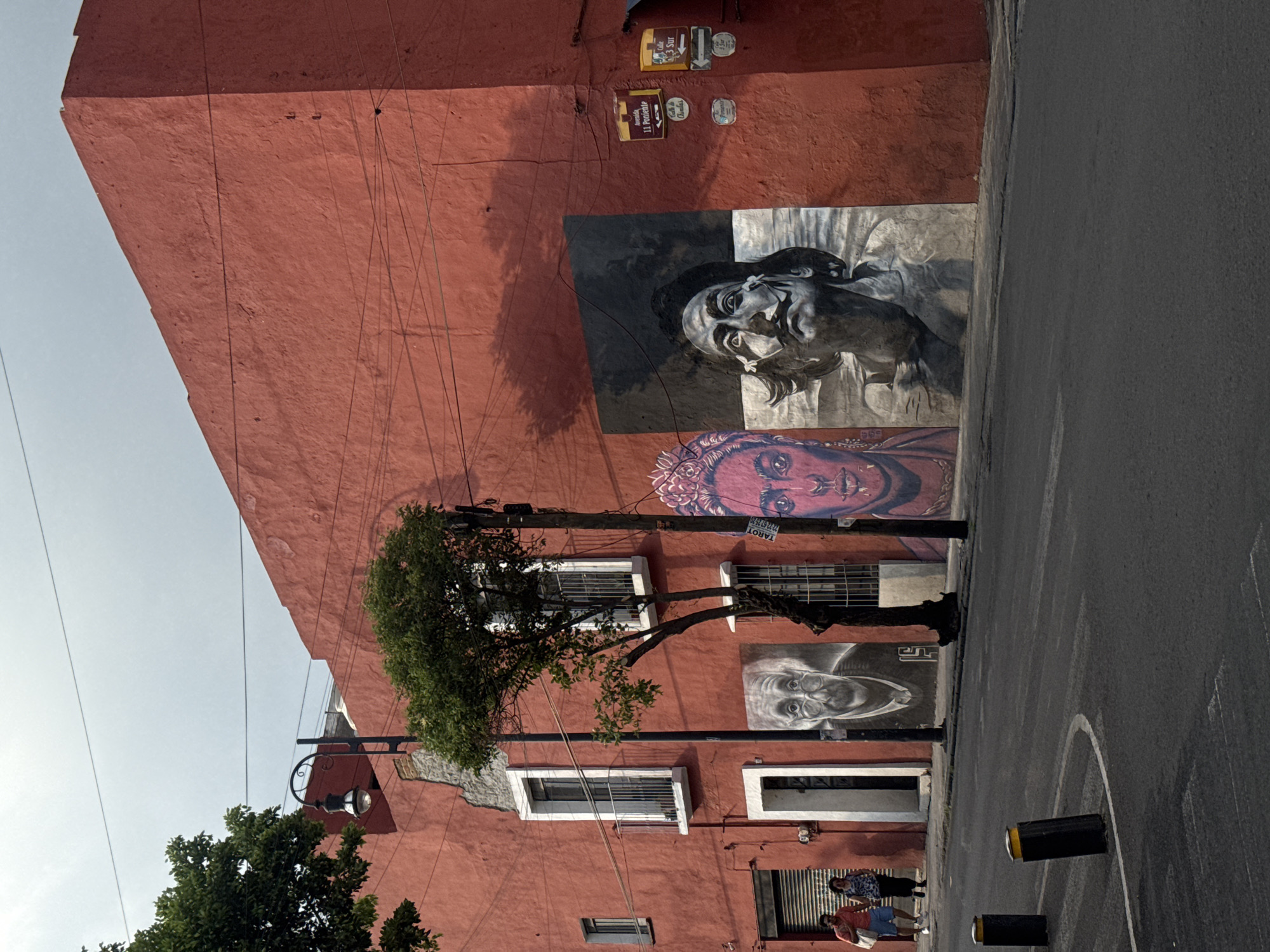 The image depicts a red building in Puebla, Mexico, adorned with three distinct murals.
From top to bottom, the murals feature:
* A grayscale portrait of a man.
* A colorized portrait with a prominent headdress.
* A grayscale portrait of an older man wearing glasses.
The building has white-framed windows. Power lines are visible against the sky. The street in front of the building is paved and marked with white lines. Two bollards are placed along the curb.
Two people are standing on the sidewalk near the building. A small tree is partially obscuring the building and the murals. Several small signs are attached to the building above the top mural.