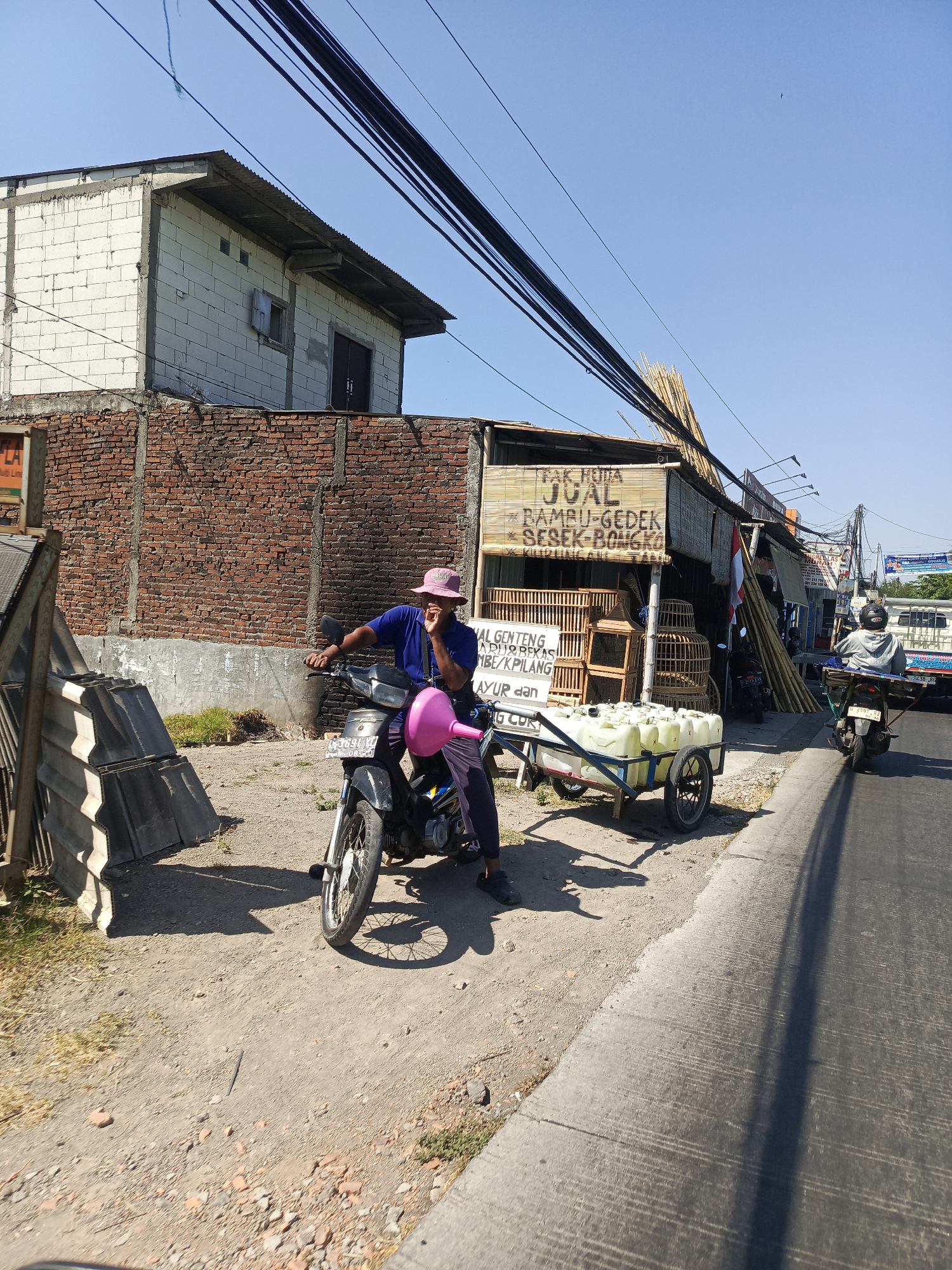 In Dukuhtengah, Indonesia, on a sunlit day, a man wearing a blue shirt and a pink hat sits astride a black motorcycle, a pink funnel attached to its side. He appears to be holding a conversation. The motorcycle is parked on a dirt road next to a sidewalk. Behind him, a vendor stall displays bamboo products and birdcages, with stacks of bamboo poles leaning against the building. The stall has signage written in Indonesian. Across the street, a white truck and another motorcycle are visible. The scene is framed by a brick building and overhead power lines.