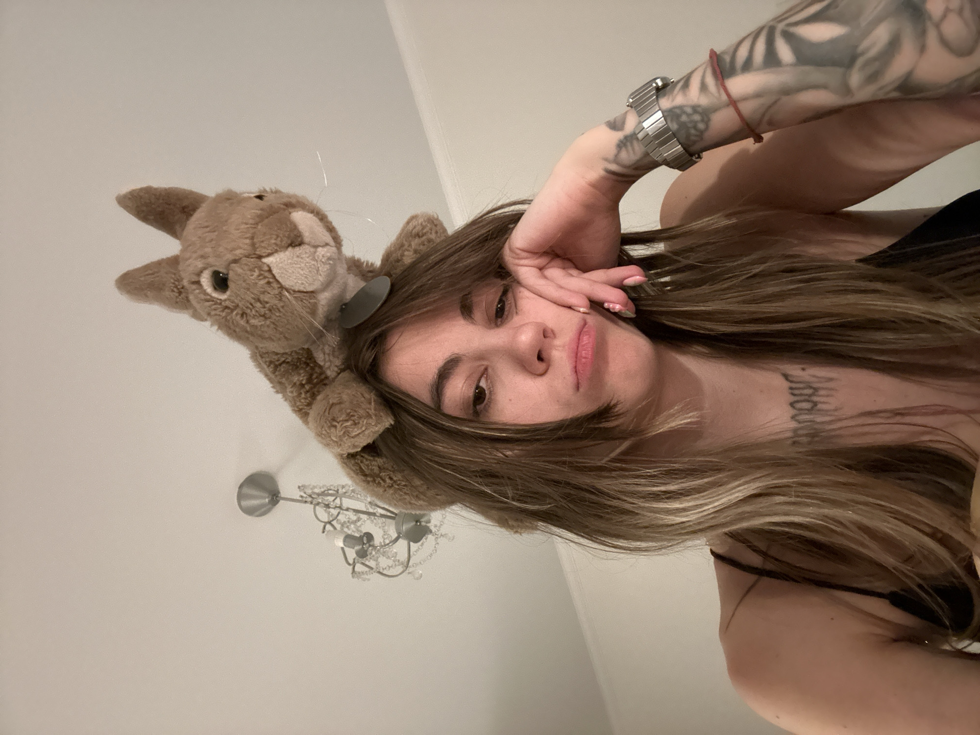 A young woman with long, light brown hair and prominent tattoos on her right arm and chest is shown in a close-up, low-angle shot. She has a bored or tired expression, with her chin resting on her hand. A stuffed squirrel toy is positioned on top of her head. Behind her, a chandelier with crystal accents hangs from the ceiling, and the walls are a neutral color. The scene appears to be indoors, possibly in a residential setting. There is no indication of a specific location in Moscow, Russia.