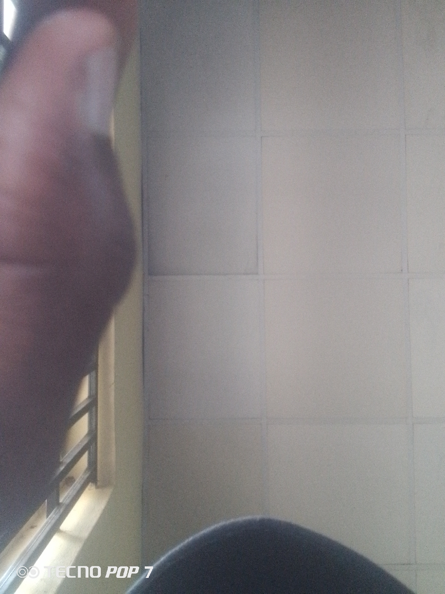 The image is a close-up, low-angle shot of a tiled wall, likely indoors, with a blurred foreground element that appears to be a person's arm or leg. A sliver of a window with horizontal bars is visible on the left, suggesting a possible view to the outside or another room. The dominant colors are muted grays and a yellowish hue from the wall behind the bars. The bottom of the frame shows a watermark that reads "TECNO POP 7." The scene is devoid of any specific identifying features that would place it within the city of Eleweran, Nigeria.