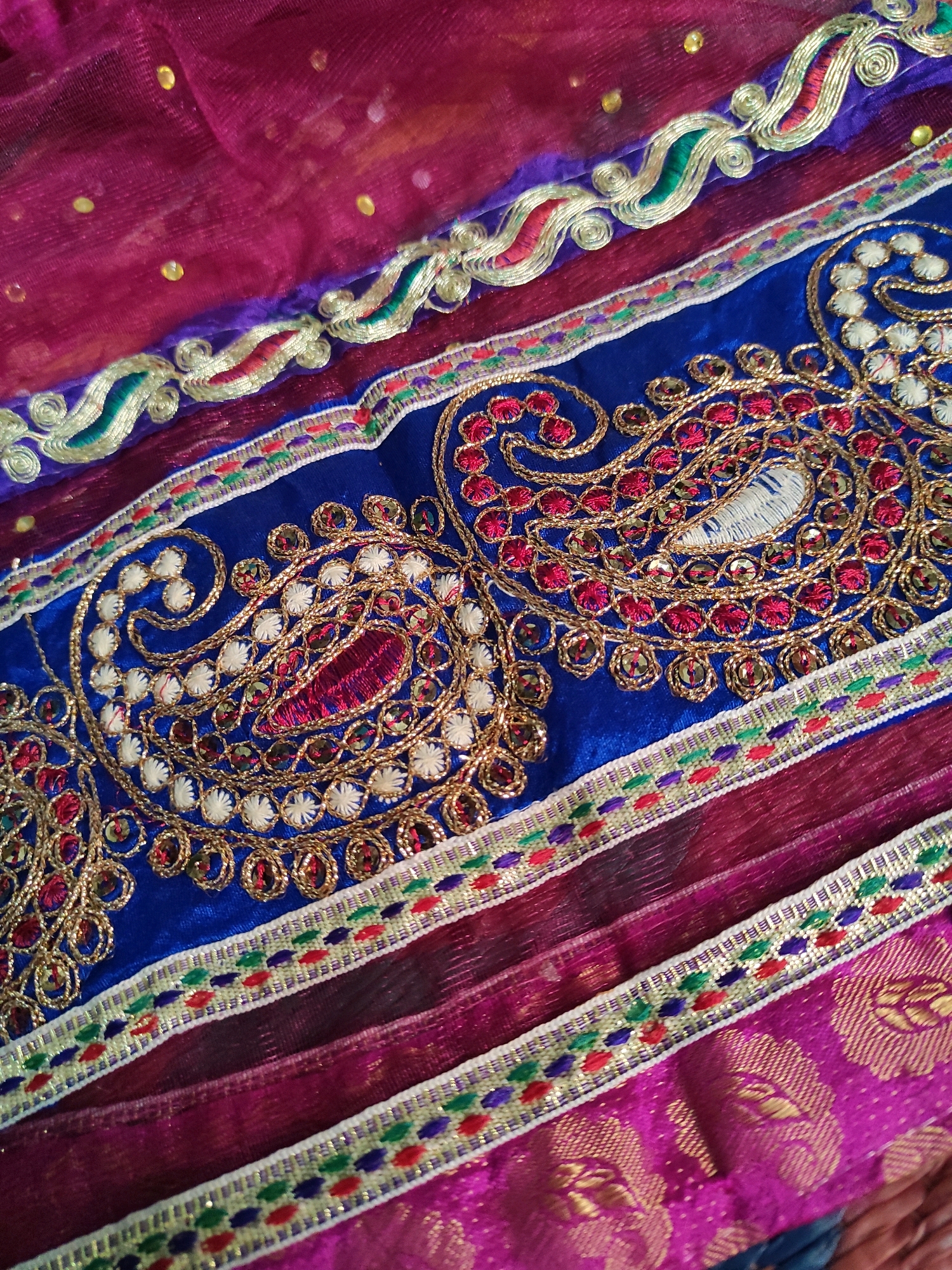 The image displays a close-up of an intricately embroidered Indian textile, likely part of a sari or dupatta. The dominant colors are vibrant magenta, deep royal blue, and gold. The fabric features elaborate paisley motifs embroidered with gold thread, accented by red and white bead-like stitching and small, reflective sequins. Decorative borders with wave-like patterns in gold, green, and red, along with bands of colorful stitched dots, frame the central blue embroidered section. The textile appears to be layered, with a sheer magenta fabric overlaid on a richer, patterned magenta silk-like material at the bottom. The lighting suggests an indoor setting, possibly a retail environment or a home. There are no people or specific landmarks visible to pinpoint a location within Hyderabad, India. The image captures the rich craftsmanship and detail of traditional Indian embroidery.