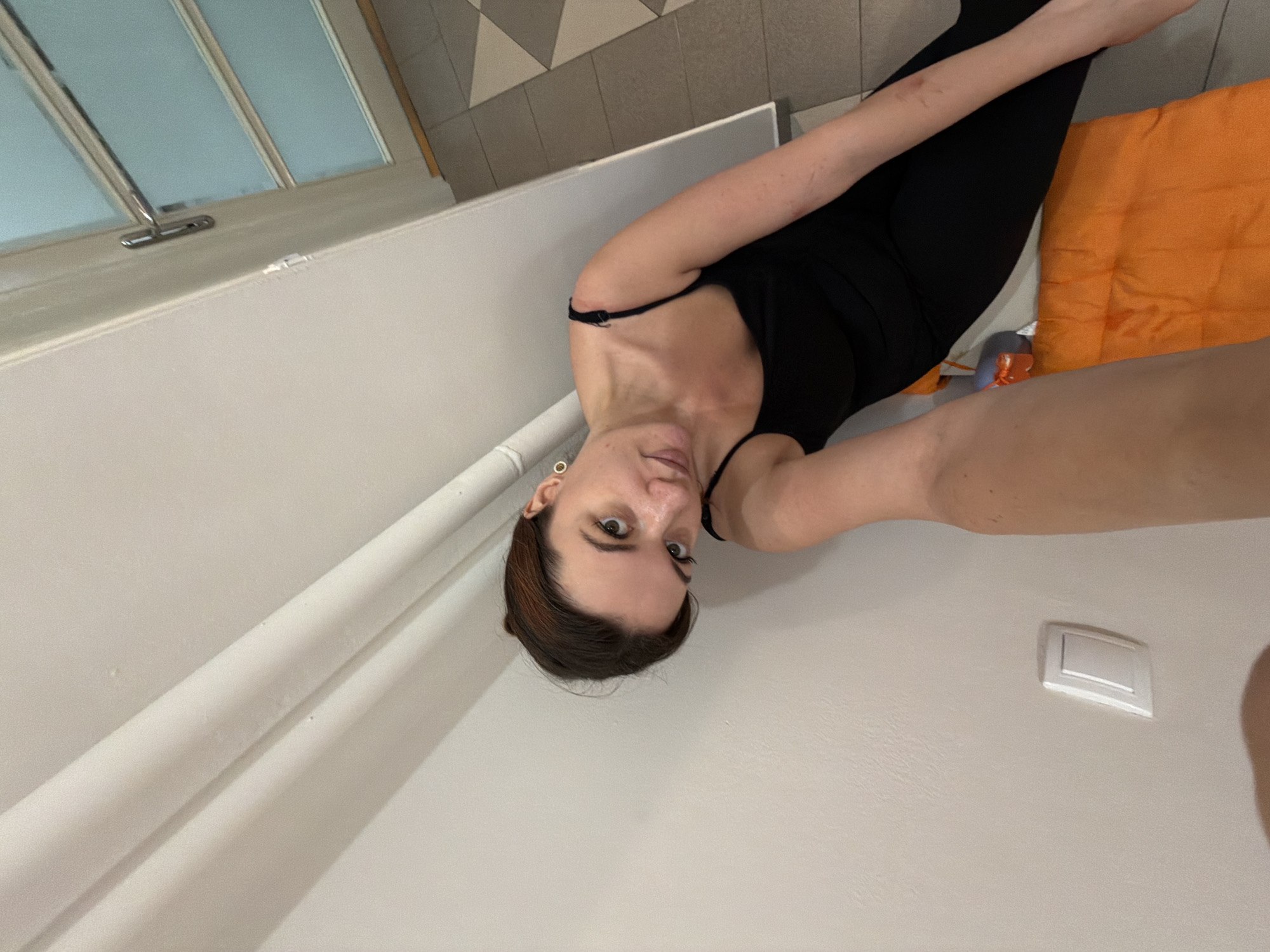 A woman in a black leotard and leggings is lying on her side in a room with a window and a light switch on the wall. An orange pillow is on the floor. The location could be in Belgrade, Serbia, but there are no clues in the image to confirm this.