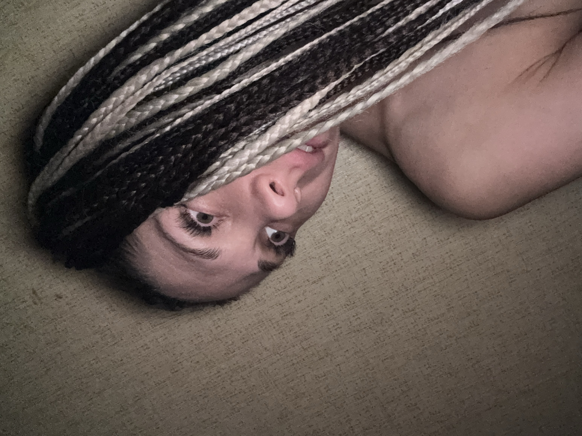 A person is lying on a textured surface, possibly a couch or bed, with their head positioned upside down relative to the viewer. The person has dark and light-colored braided hair that partially covers their face. Their eyes are open, and they are looking upwards. A portion of their shoulder is visible. The location is unknown.