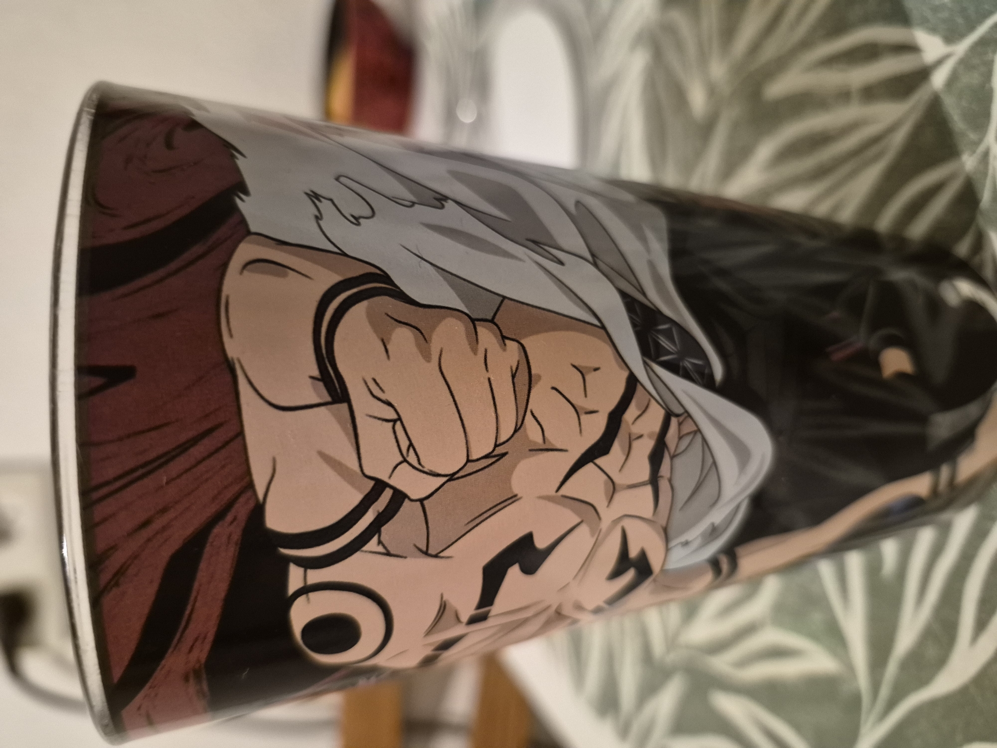 A close-up, slightly angled view shows a glass with an anime-style illustration of a muscular character with dark hair and a light-colored shirt. The character's fist is prominent in the foreground, with visible muscle definition and black markings on the arms and torso. The glass is placed on a table with a patterned green tablecloth. In the blurred background, a red bowl and white plates are visible. The scene is indoors, with the lighting suggesting it is daytime.