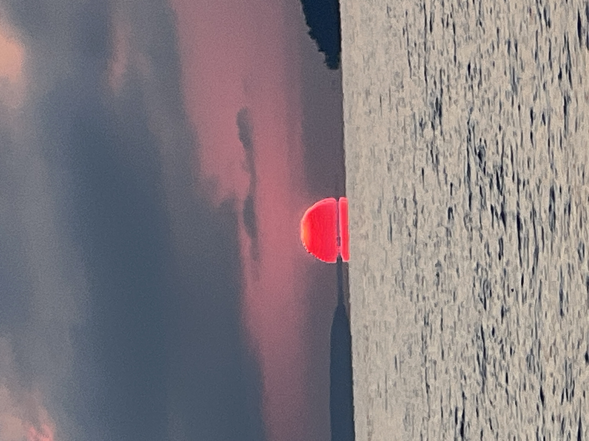 This is a close-up, eye-level shot of a sunset over a body of water. The sun, a vibrant red orb, is partially submerged at the horizon, casting a colorful reflection on the water's surface. The sky is a gradient of dark gray clouds at the top, transitioning to shades of pink and red closer to the horizon. The water itself is calm, with subtle ripples creating a textured effect. In the distance, the silhouette of a landmass, likely a hill or a small mountain, is visible on the right side of the frame, with a darker, flatter landmass on the left. The scene is devoid of any people or man-made objects, focusing solely on the natural beauty of the sunset.