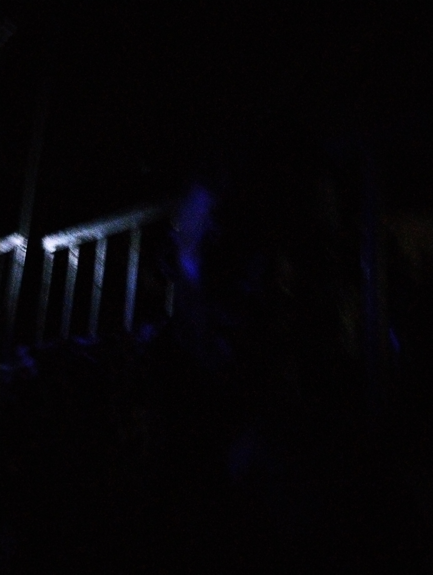 The image shows a dark scene. In the left portion of the frame, there are a few vertical bars of a railing illuminated by an unseen light source. To the right, there is a vague blue-tinged form, possibly a person or object. The environment is very dark, obscuring any background details that would locate this scene in Fegge, Nigeria.