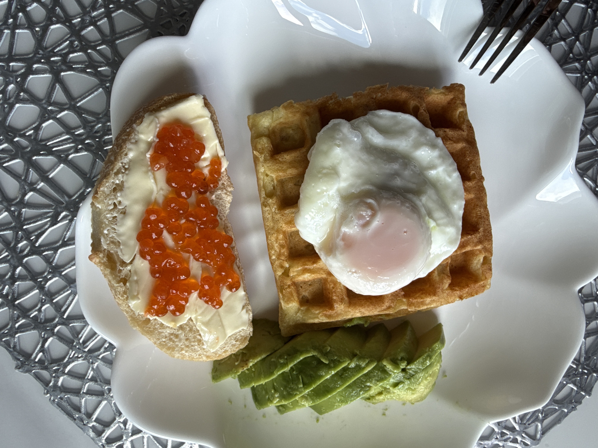 This is a top-down, close-up, eye-level shot of a breakfast plate set on a silver placemat. The plate is white and has a scalloped edge. On the plate, from top to bottom, left to right, are: a slice of bread spread with butter and topped with red caviar, several slices of avocado, a waffle topped with a poached egg, and a fork on the right edge of the plate. The background is the silver placemat, which has a textured, net-like pattern. The overall lighting is bright, with some slight reflections on the plate. The image does not provide any information about the city of Khimki, Russia.