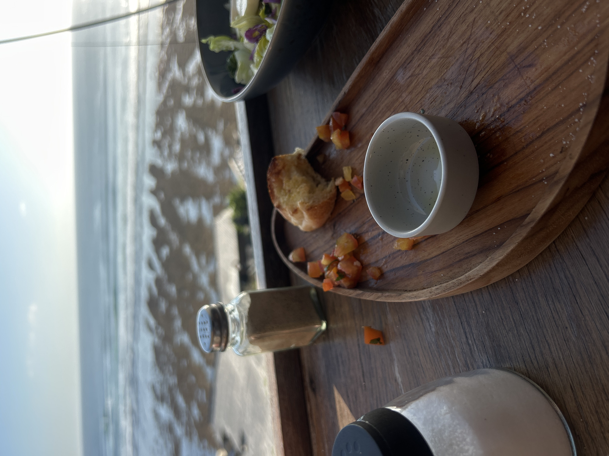 A meal is laid out on a wooden table in front of a large window overlooking a beach and the ocean. In the foreground, a wooden tray holds a small white bowl with a clear liquid, a piece of bread, and scattered diced tomatoes. To the left, two salt shakers are visible: one tall glass shaker filled with white salt and a smaller glass shaker with a metallic top filled with brown seasoning. In the background, a dark bowl contains a salad with lettuce, purple cabbage, and possibly white cheese. The ocean stretches to the horizon under a bright sky, and the wet sand of the beach is visible in the middle ground. The scene appears to be set in Kuta Selatan, Indonesia, likely at a restaurant or cafe with a beach view.