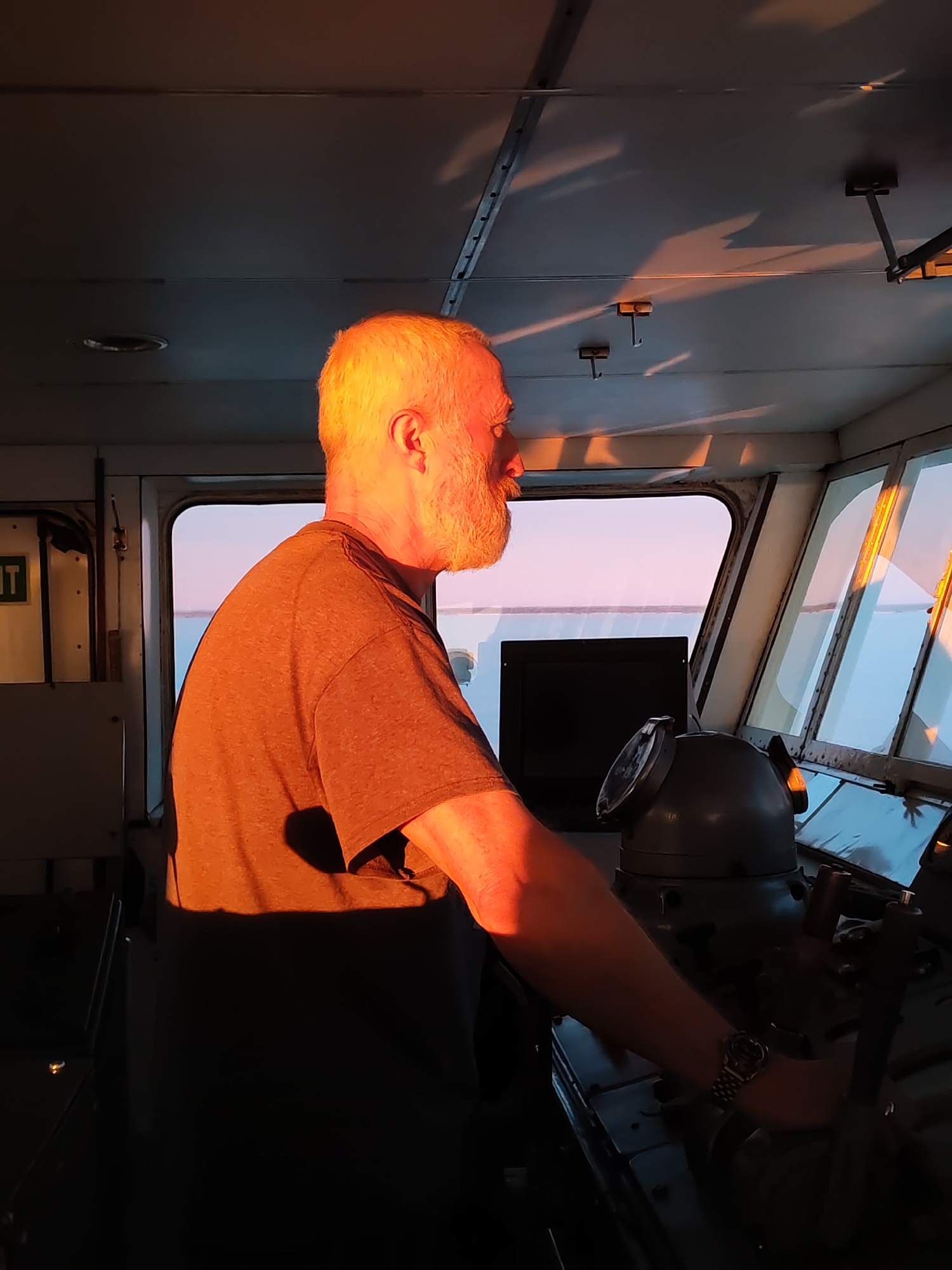 A middle-aged man with white hair and a beard is at the helm of a ship. He is wearing a gray t-shirt and a watch. His right arm is on the steering wheel. He looks out the window. The bridge of the ship is metallic with a dark gray control panel. Sunlight streams through the windows and illuminates the man and the bridge. The man is not in Bereg, Russia.