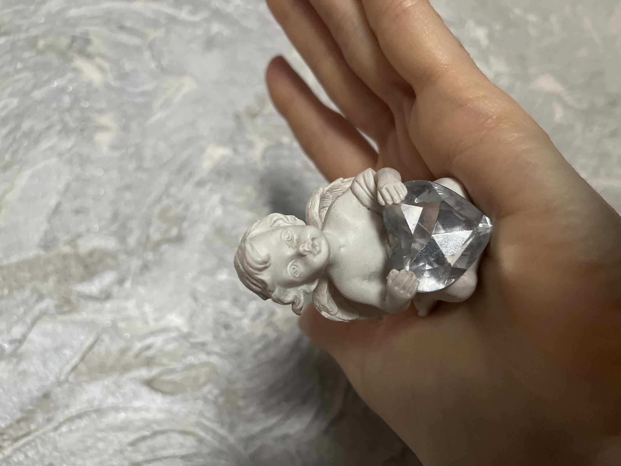 A hand holds a small, white angel figurine clutching a large, clear crystal. The background is a textured, gray-white surface. The image does not provide enough information to determine the location, including the city of Krasnodar, Russia.