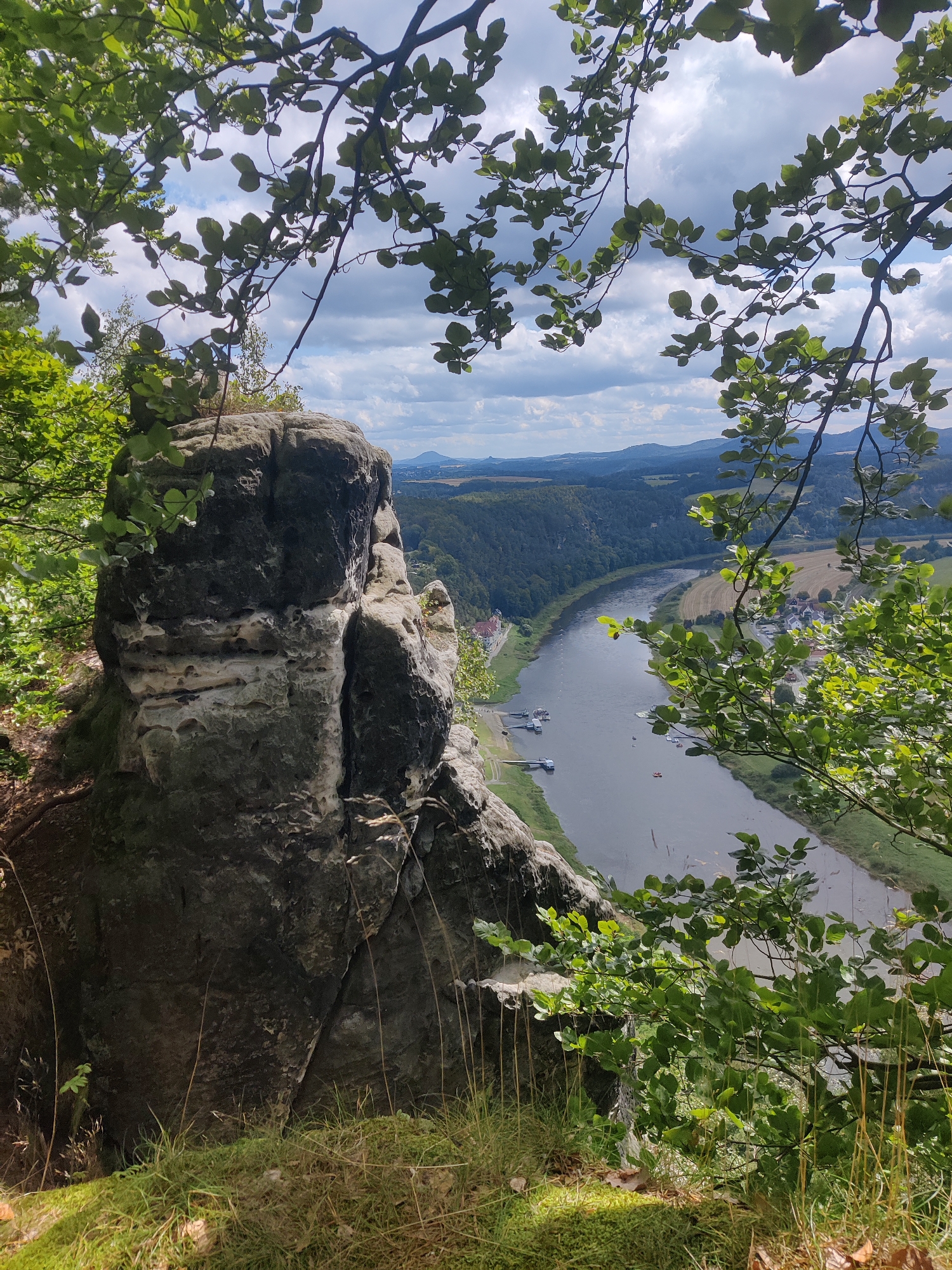 The image captures a scenic view from a rocky outcrop overlooking the Elbe River valley near Lohmen, Germany. In the foreground, a large, weathered rock formation stands prominently, partially obscured by lush green foliage and branches of trees. The rock displays distinct layers and textures, with some areas appearing lighter and eroded, while others are darker and more solid.
Beyond the rock, the Elbe River meanders through a valley. Several boats, including what appear to be tour boats and smaller vessels, are visible on the water, some docked along the riverbank. A small town or village is situated on the right bank of the river, with houses and buildings nestled amidst green fields and trees. The landscape is characterized by rolling hills and distant mountains, all under a partly cloudy sky. The vegetation is dense and verdant, suggesting a temperate climate. The overall impression is one of natural beauty and tranquility, with hints of human activity integrated into the landscape.