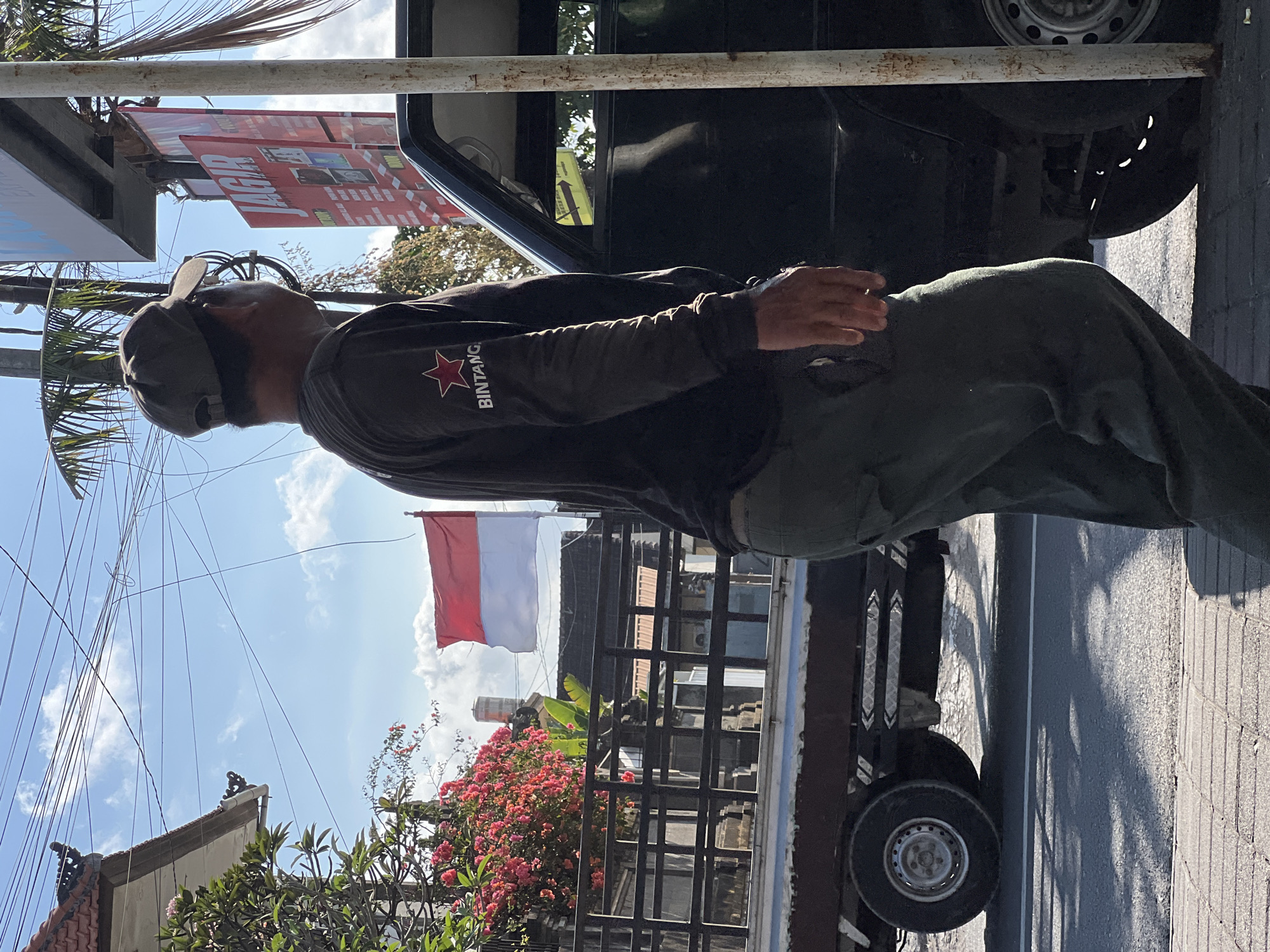 An individual wearing a dark long-sleeved shirt with a red star logo and the word "BINTANG" is seen from the side, standing near a black vehicle. They are also wearing a dark baseball cap and olive green pants. In the background, an Indonesian flag is visible, along with a truck, buildings, trees, power lines, and a street. The scene appears to be a street in Denpasar, Indonesia, characterized by typical urban elements and sunny weather.