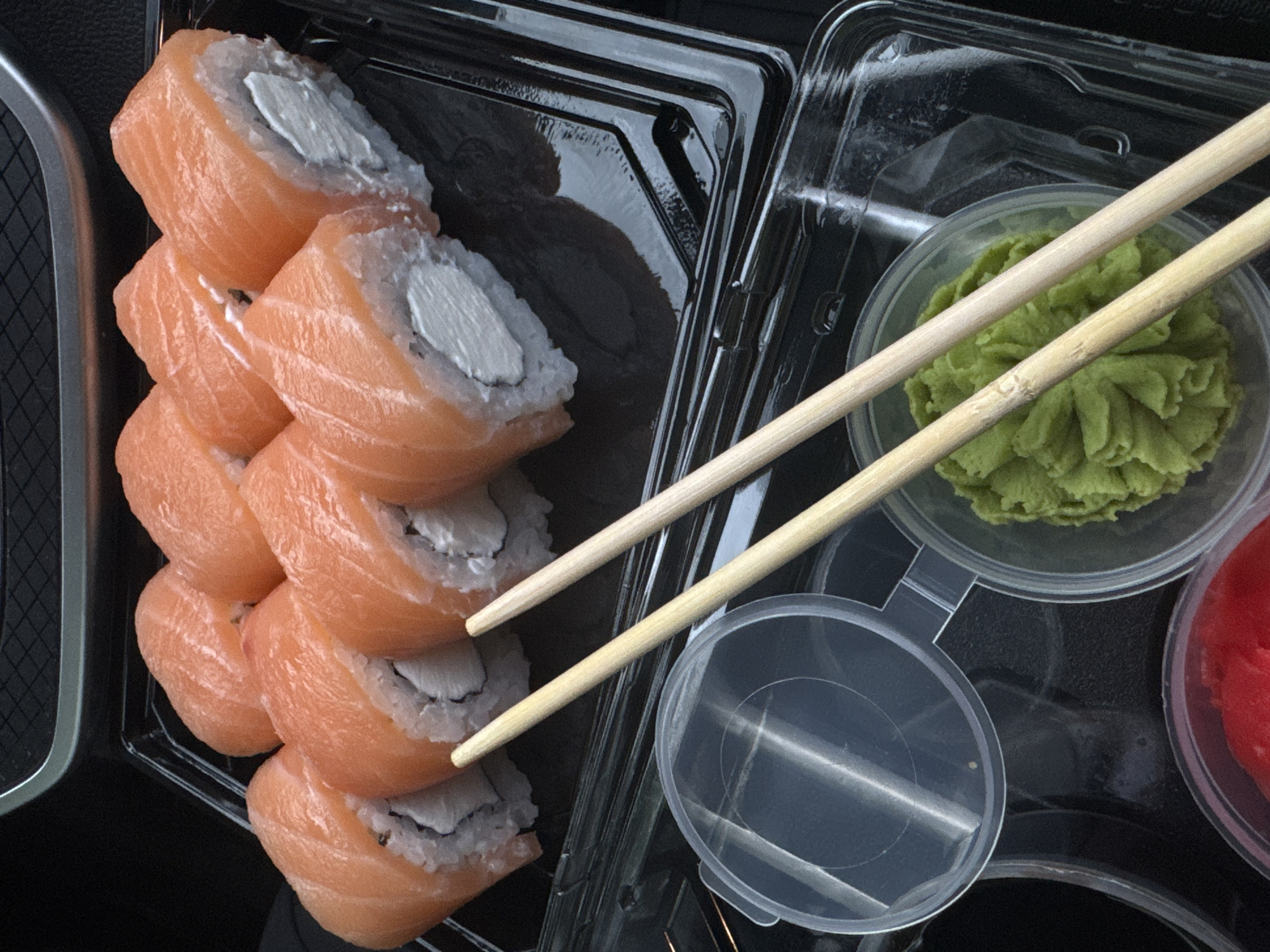 A close-up, overhead shot showcases a black plastic takeout container filled with Philadelphia rolls, a type of sushi. The rolls are arranged in two rows, with the front row showing six pieces and the back row partially visible. Each roll is topped with a vibrant orange slice of salmon, enclosing a creamy white filling, likely cream cheese, and white rice. Two light-colored wooden chopsticks are placed diagonally across the lower half of the sushi container, resting on top of the rolls.
Below the sushi, a clear plastic container holds a dollop of bright green wasabi, and another partially obscured container appears to contain pickled ginger. A clear plastic lid is also visible in the lower left corner of the frame. The background is dark and appears to be the interior of a vehicle, with a textured dark grey surface in the upper portion of the image. The scene is set in Mikhaylovsk, Russia.
