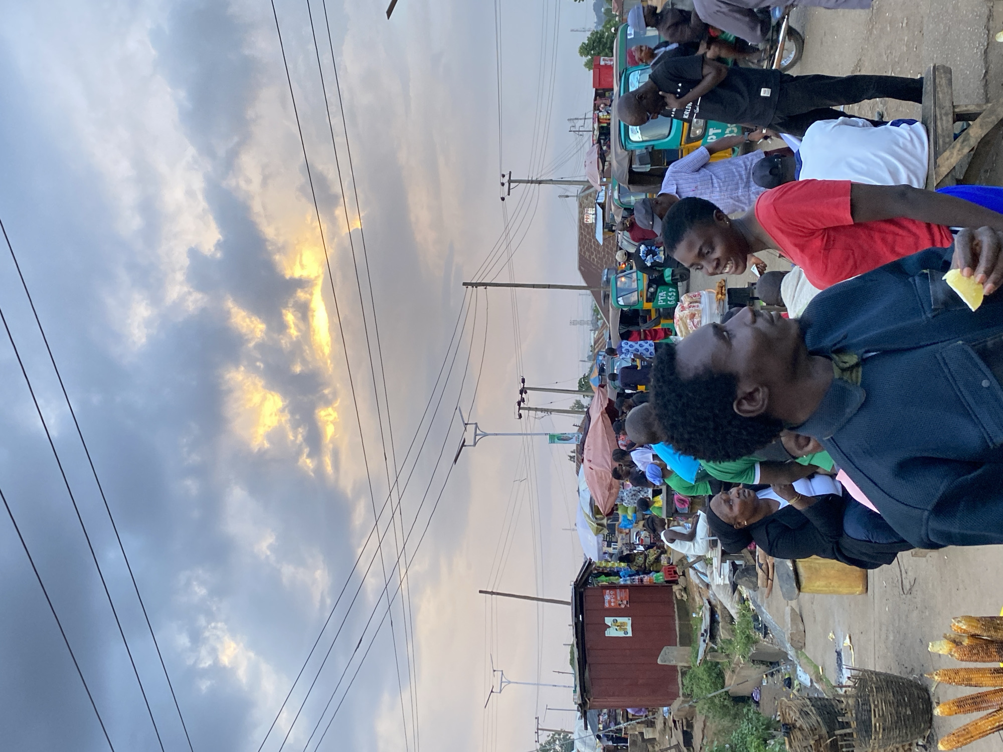 The image depicts a bustling outdoor market scene in Bukuru, Nigeria, under a dramatic sky with patches of sunlight breaking through heavy clouds. In the foreground, a man in a dark blue jacket holds a slice of lemon. Beside him, a woman in a red t-shirt smiles. Behind them, a crowd of people are engaged in various activities.
Several three-wheeled auto-rickshaws, commonly known as "Keke Napep" in Nigeria, are parked, some appearing to be for hire. One rickshaw has the license plate "PTA 6657". Stalls with umbrellas and awnings are set up, suggesting vendors are selling goods. Items like corn on the cob are visible in baskets. Buildings and makeshift structures line the background. Overhead power lines crisscross the sky. The overall impression is one of a lively commercial hub.