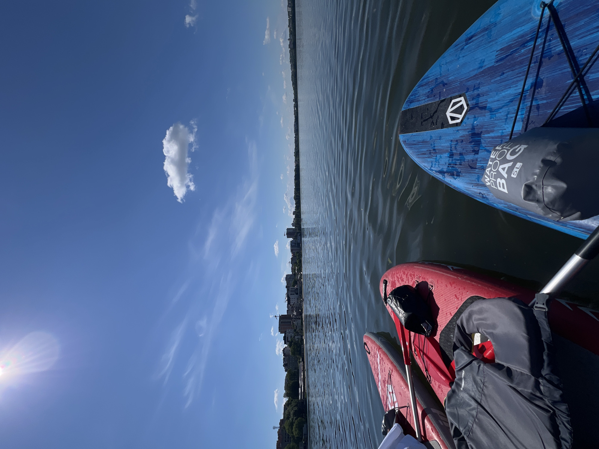 The image depicts a serene scene on the Ternopil pond in Ternopil, Ukraine, on a bright, sunny day. The perspective is from a paddleboard, looking out over the calm water towards the city skyline. Two red paddleboards are visible in the foreground, with a grey life jacket and a black bag resting on one of them. A blue and black paddleboard, also equipped with a grey waterproof bag, is closer to the viewer and positioned to the right. The water is rippled, reflecting the clear blue sky with scattered wispy clouds and the bright sun. In the distance, the urban landscape of Ternopil is discernible, characterized by various buildings, trees, and a hint of greenery lining the shore. The overall atmosphere is peaceful and recreational, suggesting an outing on the water.