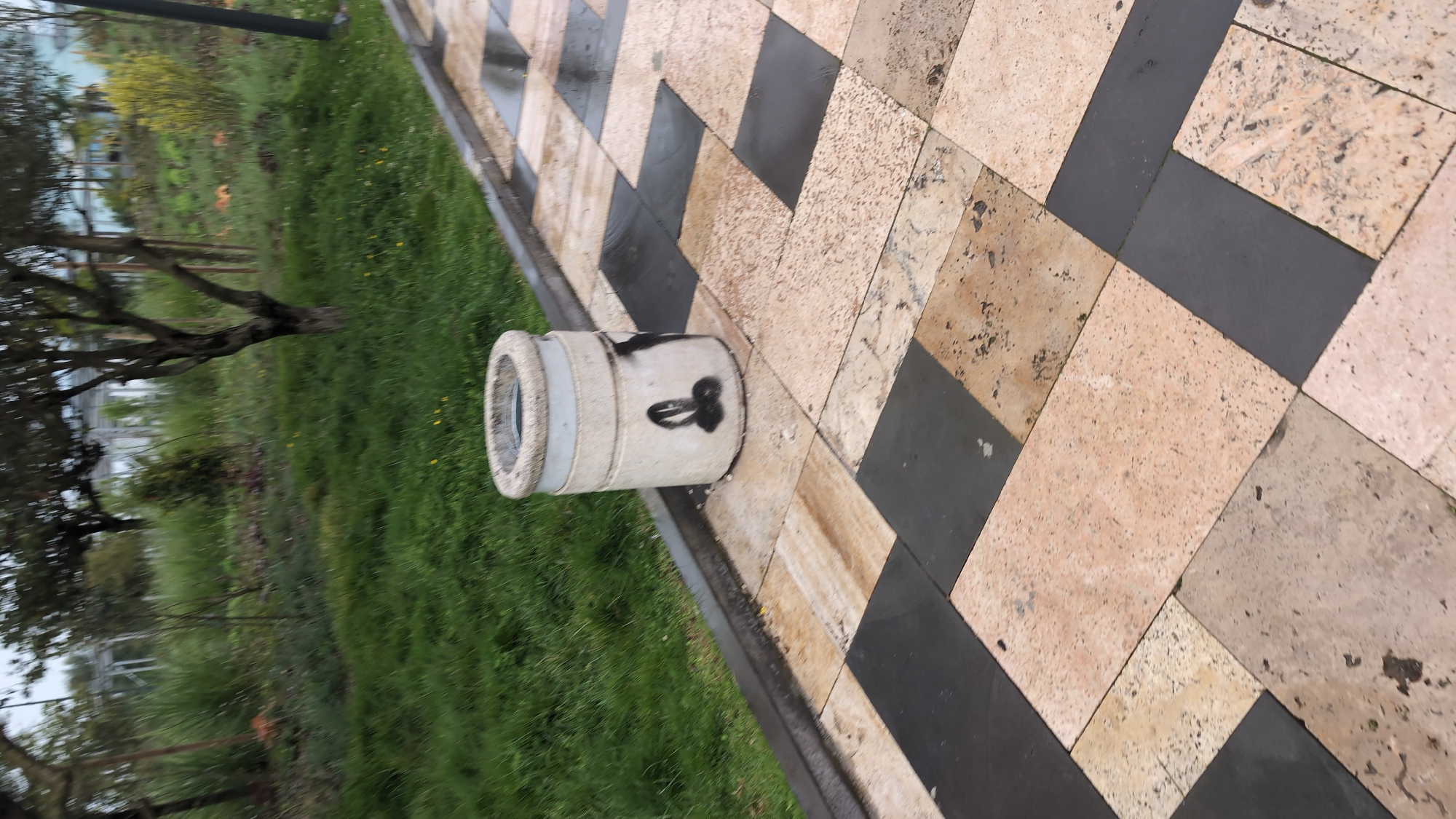 A photograph shows a cylindrical concrete bollard with graffiti on a paved pathway in Batumi, Georgia. The bollard is white with black markings. The pathway is made of alternating black and light brown stone tiles. To the left of the bollard and pathway is a strip of green grass and vegetation. In the background, trees and a building are visible. The image is taken at an angle.