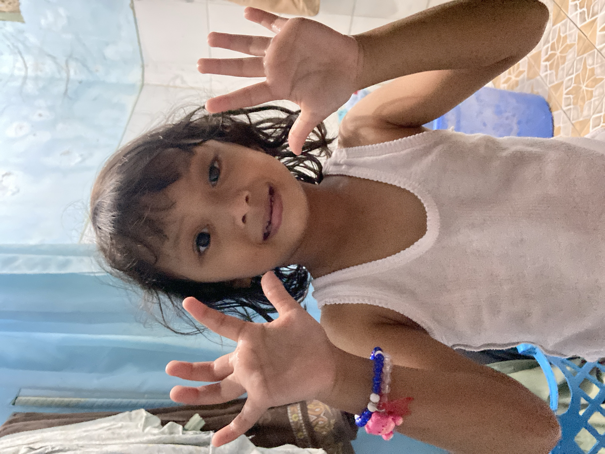 A young girl with dark hair is facing the camera and holding up both hands, fingers spread, in front of her. She is wearing a white sleeveless top and a beaded bracelet with a pink charm on her right wrist. The background consists of a wall with blue curtains or fabric on the left side, and a tiled wall with a blue plastic chair on the right side. The location is a residential interior in Gelam, Indonesia.