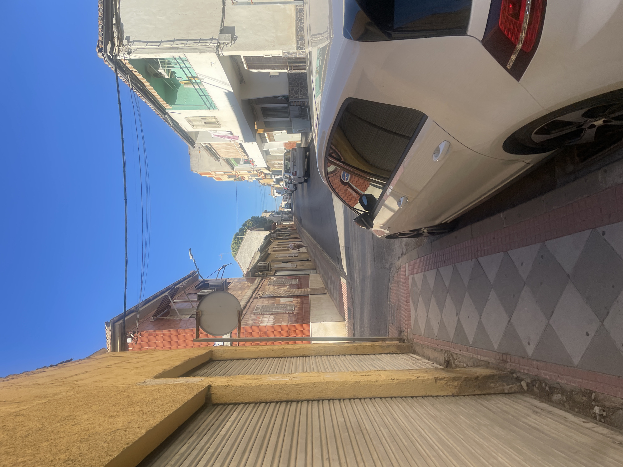 The image depicts a street scene in Atarfe, Spain, characterized by a clear blue sky and residential buildings lining a narrow road. In the foreground, a white car is parked on the right side of the street, its side and rear visible. To the left, a building with a yellow-colored facade and a closed metal shutter occupies a significant portion of the frame.
The street itself is paved with a checkered pattern of gray and light gray tiles on the sidewalk, bordered by red bricks. The road ahead curves slightly and is lined with more buildings on both sides, displaying various architectural styles and colors. Some buildings have balconies with awnings, and there are glimpses of parked cars further down the street. A single person is walking away from the camera in the distance, on the sidewalk on the right side of the road. Electrical wires are strung across the sky above the buildings. The overall impression is a quiet residential street bathed in sunlight.