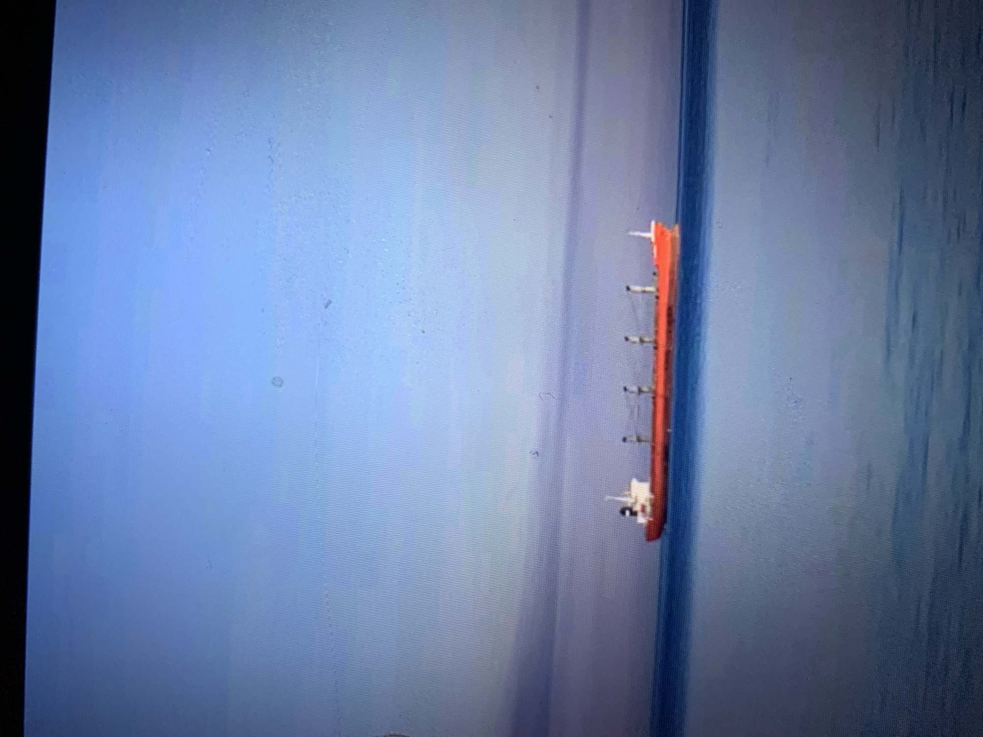 A cargo ship, predominantly bright red with a white superstructure, is sailing on a calm body of water under a pale blue sky. The ship has several tall, dark-colored funnels and a prominent white mast at its stern. The water is a deep blue and appears still, with subtle ripples on the surface. The horizon is visible as a soft, blurry line where the water meets the sky, suggesting a vast expanse of sea or ocean. The scene is serene and devoid of any human activity or other vessels, creating a sense of isolation and tranquility. The image does not provide any specific details that would allow for an accurate location within the city of Lymanka, Ukraine.