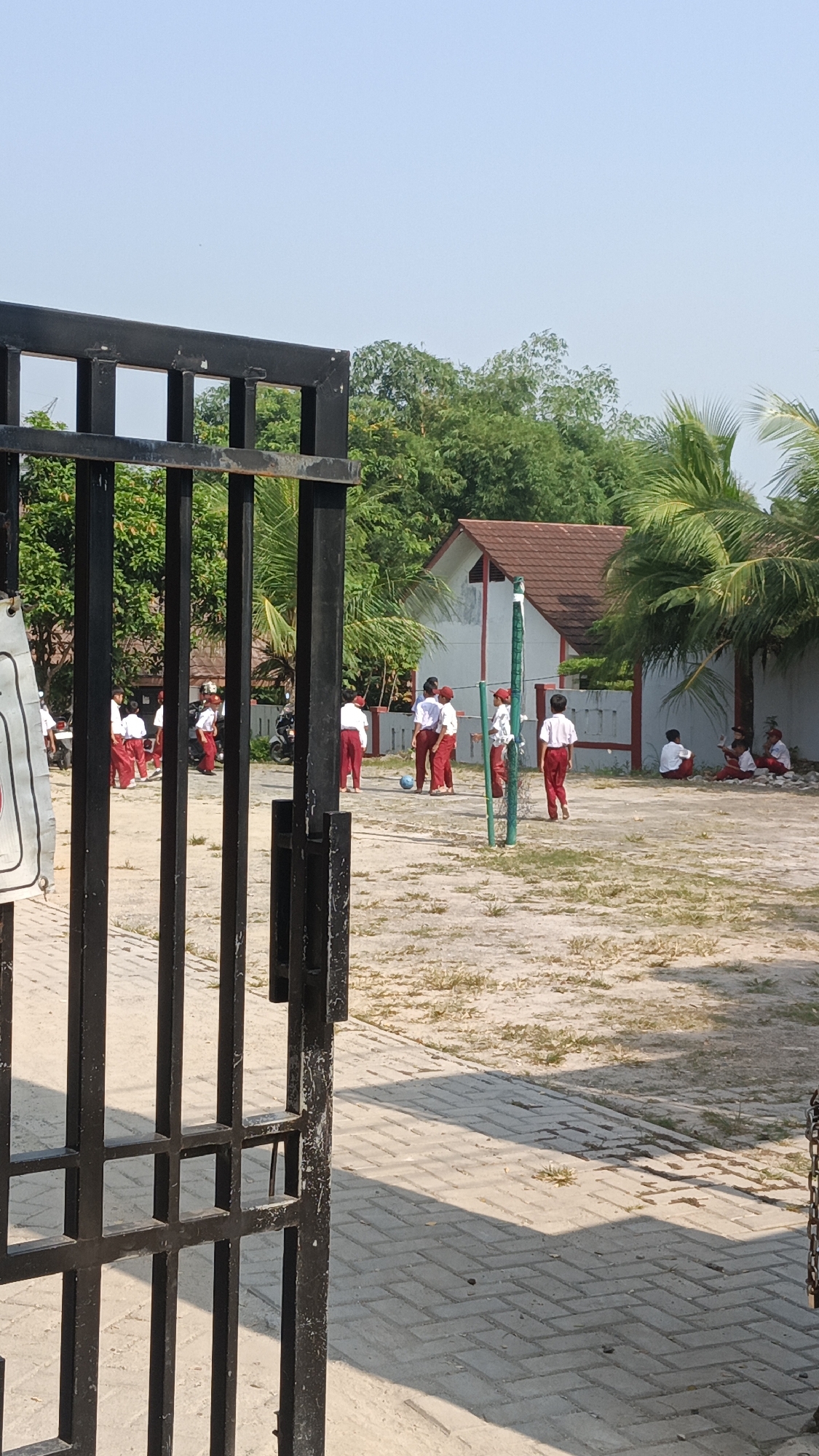 The image depicts an outdoor scene in Pasir Bolang, Indonesia, likely at a school or community area. A black metal gate is in the foreground, partially obscuring the view of a sandy and paved courtyard. Several children, dressed in white shirts and red pants or shorts, are present. Some are playing with a blue ball, while others are standing or sitting. In the background, there is a building with a brown roof and white walls, surrounded by trees and palm trees. A volleyball net is also visible. The weather appears sunny with a clear sky.