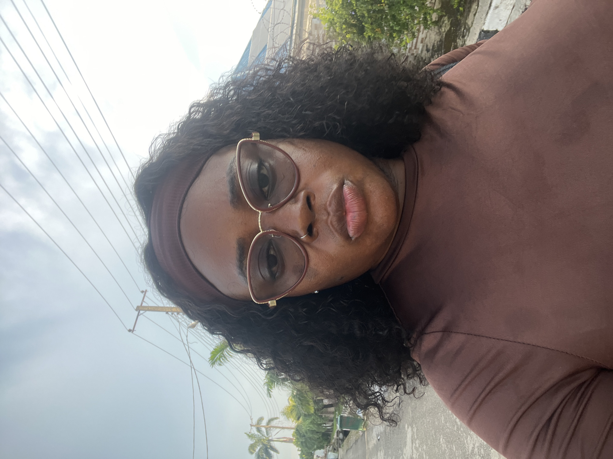 A medium shot photograph captures a Black woman from the shoulders up, looking directly at the camera. She has dark, curly hair, a dark brown headband, and is wearing large, cat-eye sunglasses with burgundy frames and gold accents. Her lips are pursed. She is wearing a dark brown, long-sleeved shirt. The background suggests an outdoor urban environment, with a paved surface, a few scattered palm trees, power lines, and some buildings and fences visible in the distance. This scene is consistent with an outdoor location within the city of Abuja, Nigeria, where such infrastructure and vegetation are commonly found.
