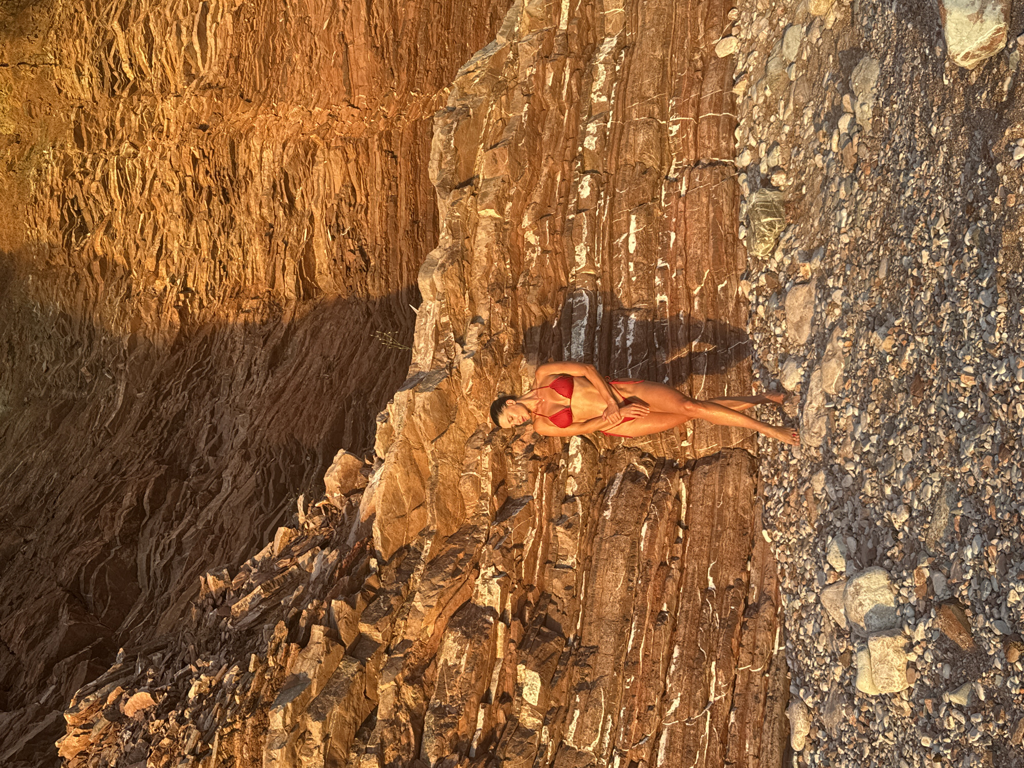 A woman stands in a red bikini on a rocky beach in front of a layered cliff face in Sveti Stefan, Montenegro. The sun, low in the sky, casts a warm, golden light on the scene, highlighting the textures of the rocks and the woman's skin. The cliff face, composed of sedimentary rock, exhibits distinct horizontal layers, some appearing to be sandstone or marl, interspersed with lighter veins of what might be calcified material. The beach is covered in a mixture of pebbles and larger, rounded rocks. The woman is posed with one leg slightly bent and crossed over the other, her head tilted down and eyes closed, appearing relaxed or contemplative. Her shadow is cast to her left on the rock face. The overall impression is one of natural beauty and tranquility.